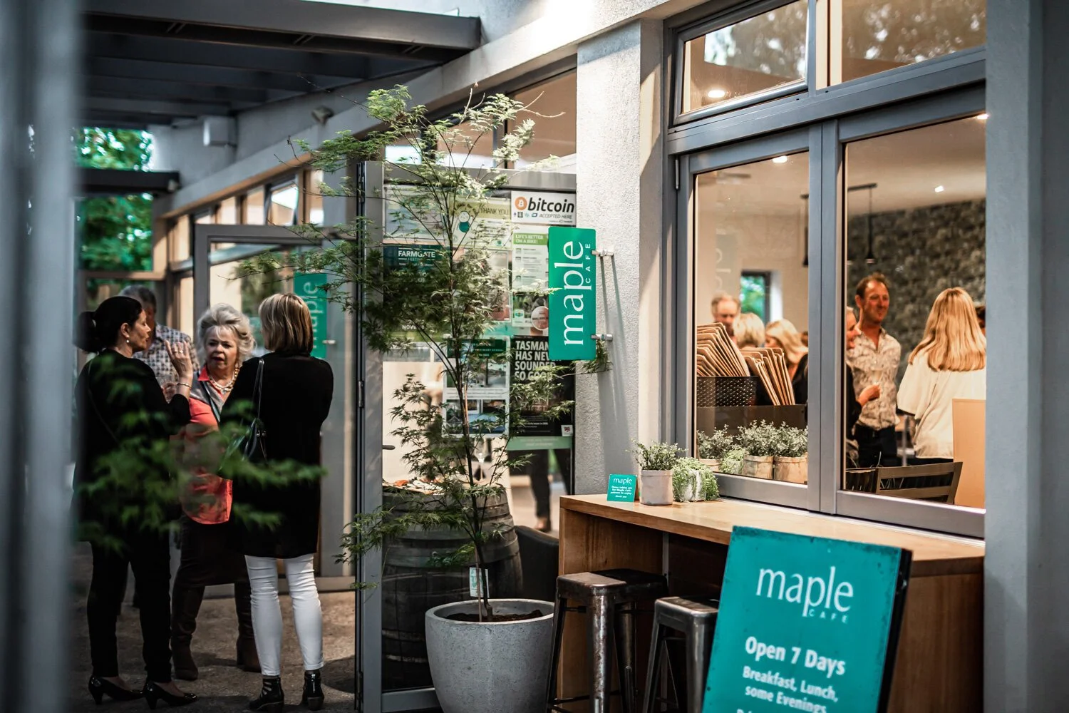 People talking outside and inside a cafe named Maple with a sign indicating it's open 7 days for breakfast, lunch, and dinner. The scene shows a small tree in a pot outside and a window with potted plants inside.