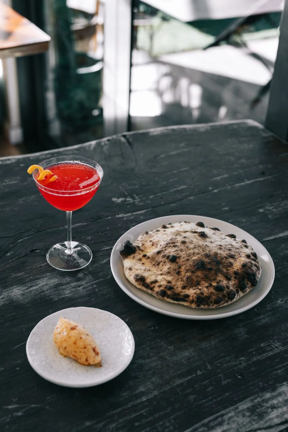 A table with a glass of red cocktail, a plate of burnt pizza bread, and a small dish with stuffed bread, in a restaurant setting.