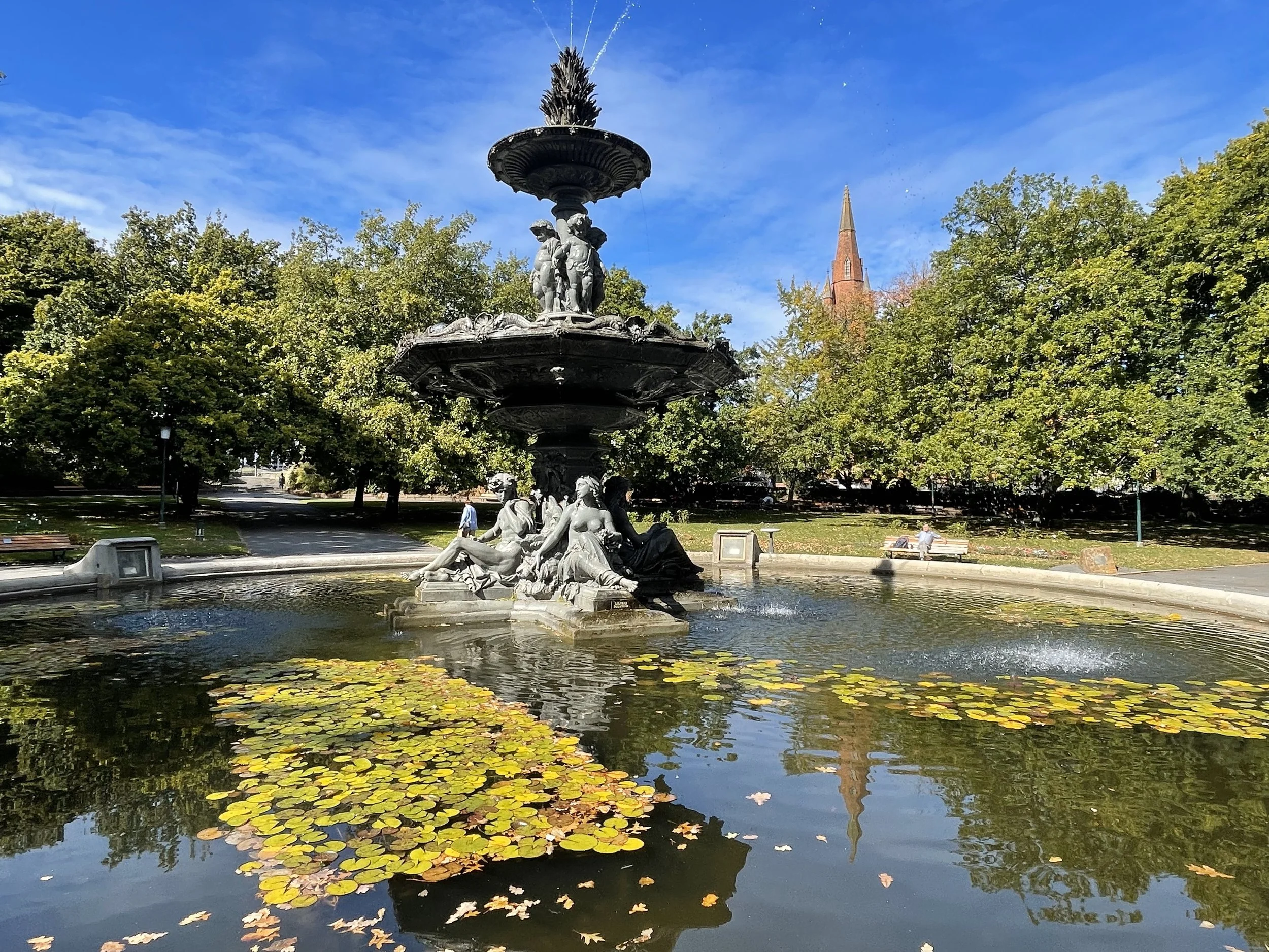 A decorative fountain in a park with water lilies in the pond, surrounded by trees and benches, under a bright blue sky with some clouds. There are people sitting on benches in the background and a church steeple is visible among the trees.