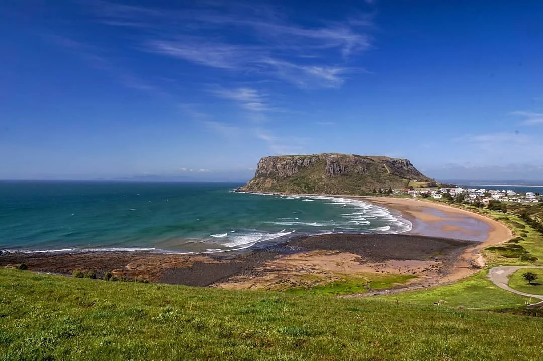 Scenic view of a coastal bay with a grassy hill in the foreground, a sandy beach curving along the shoreline, a rocky promontory in the middle distance, and a small town with white buildings and a distant ocean under a blue sky with wispy clouds.