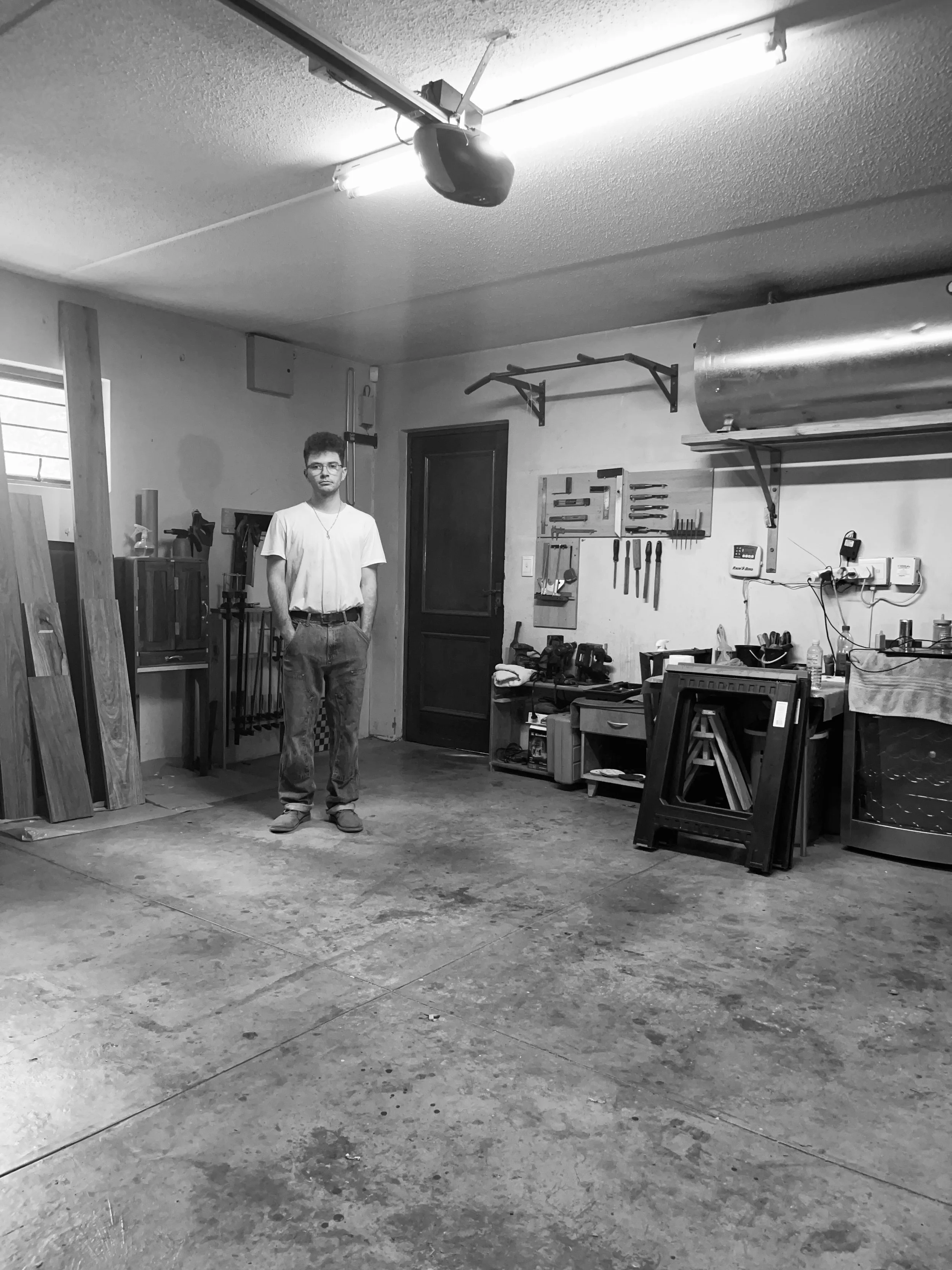 Black and white photo of a young man standing in a workshop with various woodwork tools and equipment on the wall and workbenches.