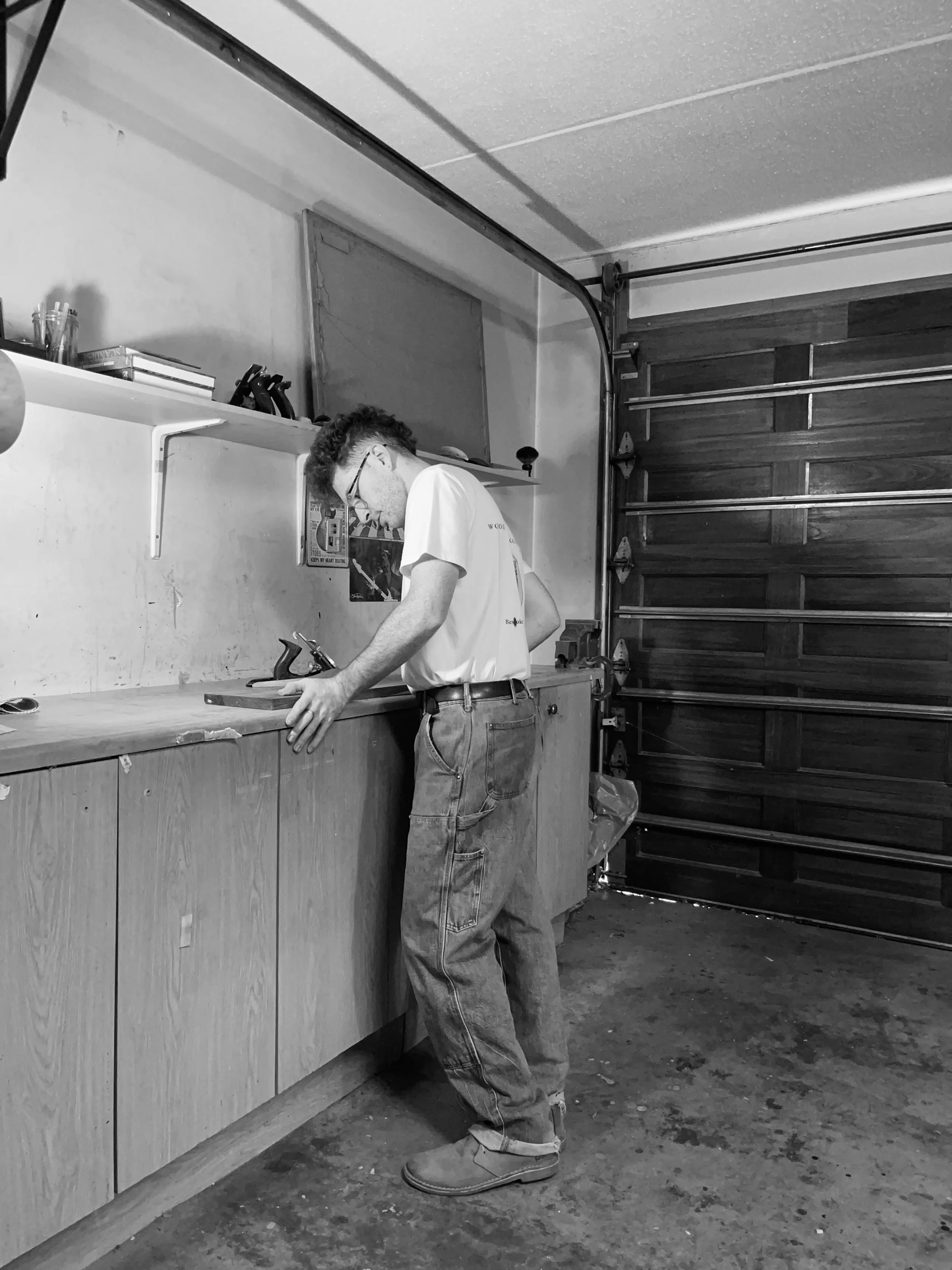 A young man working in a workshop, standing at a workbench, with woodworking tools and materials around him.