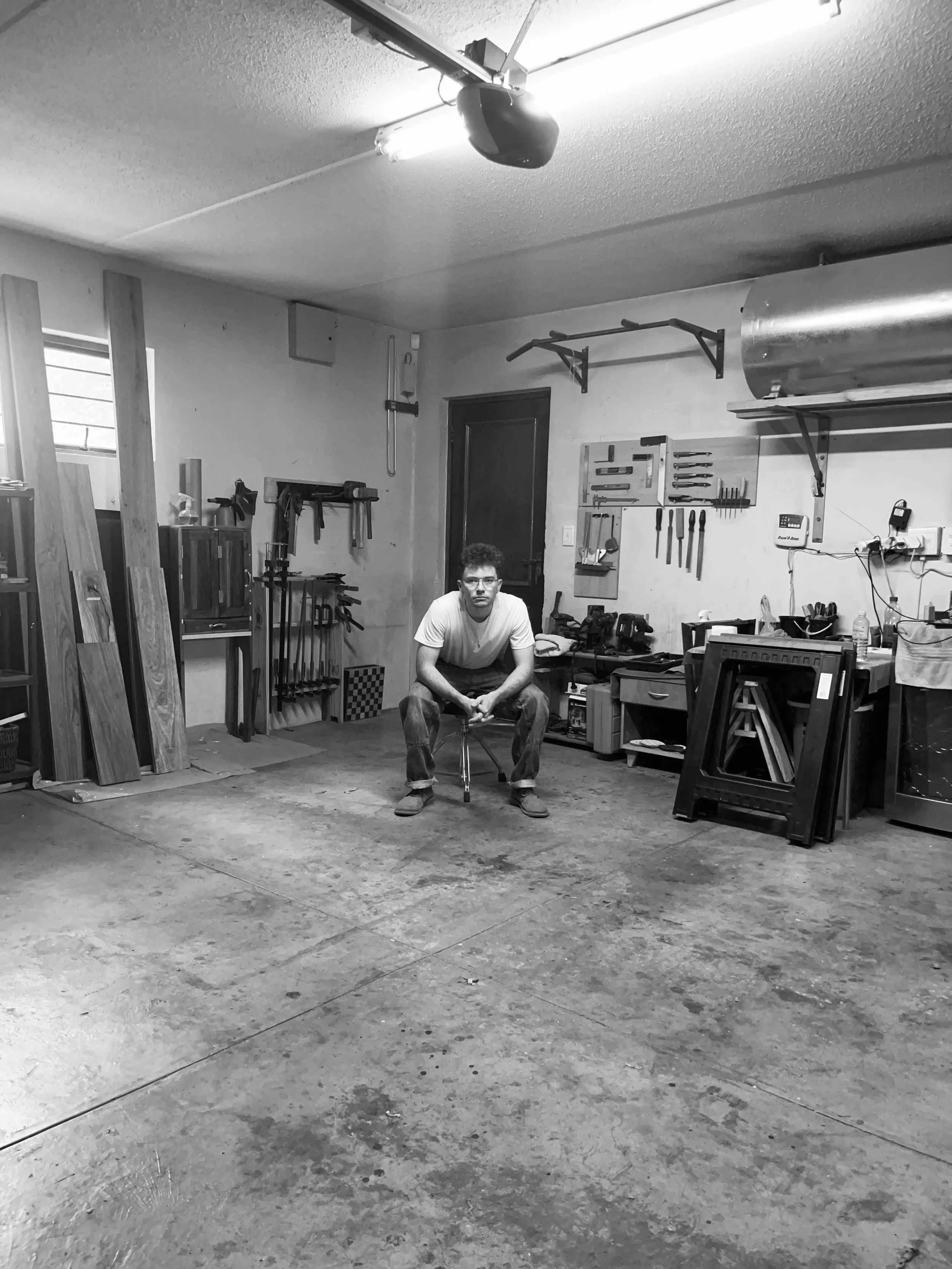 A man sitting on a stool in a woodworking workshop, surrounded by tools and lumber.
