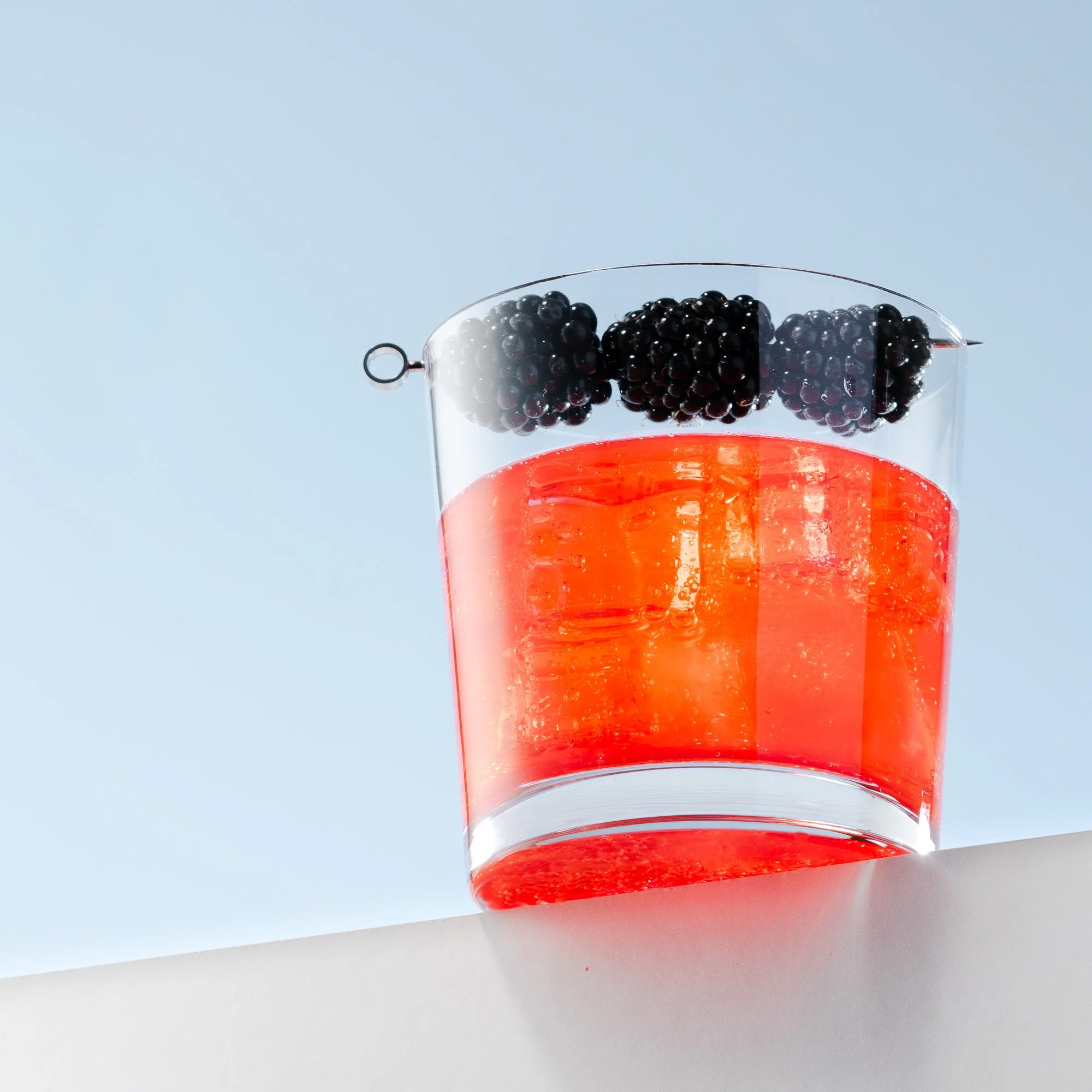 A clear glass contains red liquid at the bottom and fresh blackberries on top, set against a light blue background.