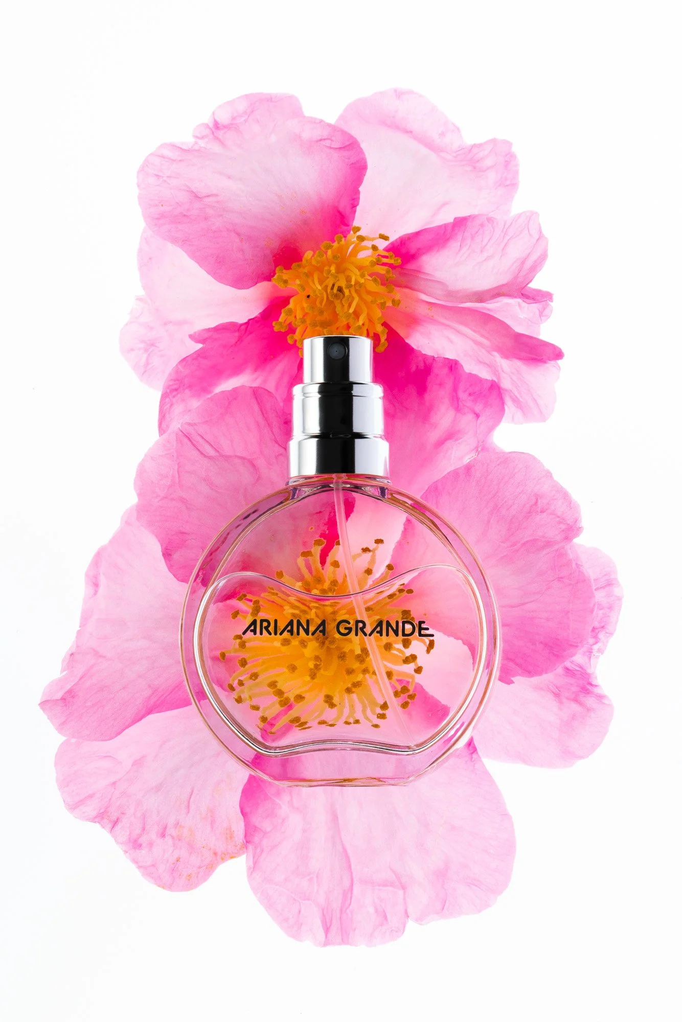 A clear perfume bottle with the label 'ARIANA GRANDE' is placed on a background of pink flower petals, and the top of the perfume bottle is surrounded by yellow flower stamen.