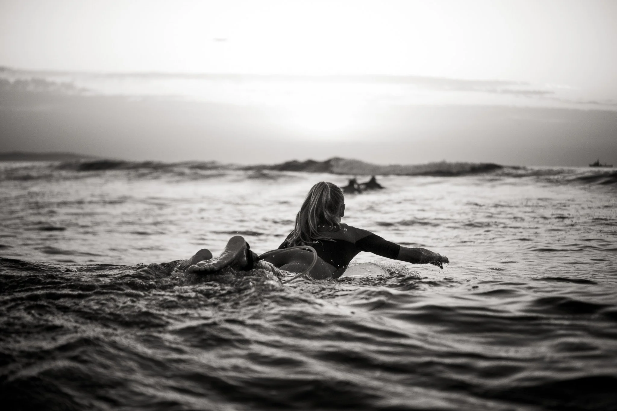 A person in a wetsuit lying on a surfboard in the ocean, with a horizon and distant boat in the background during sunset or sunrise.