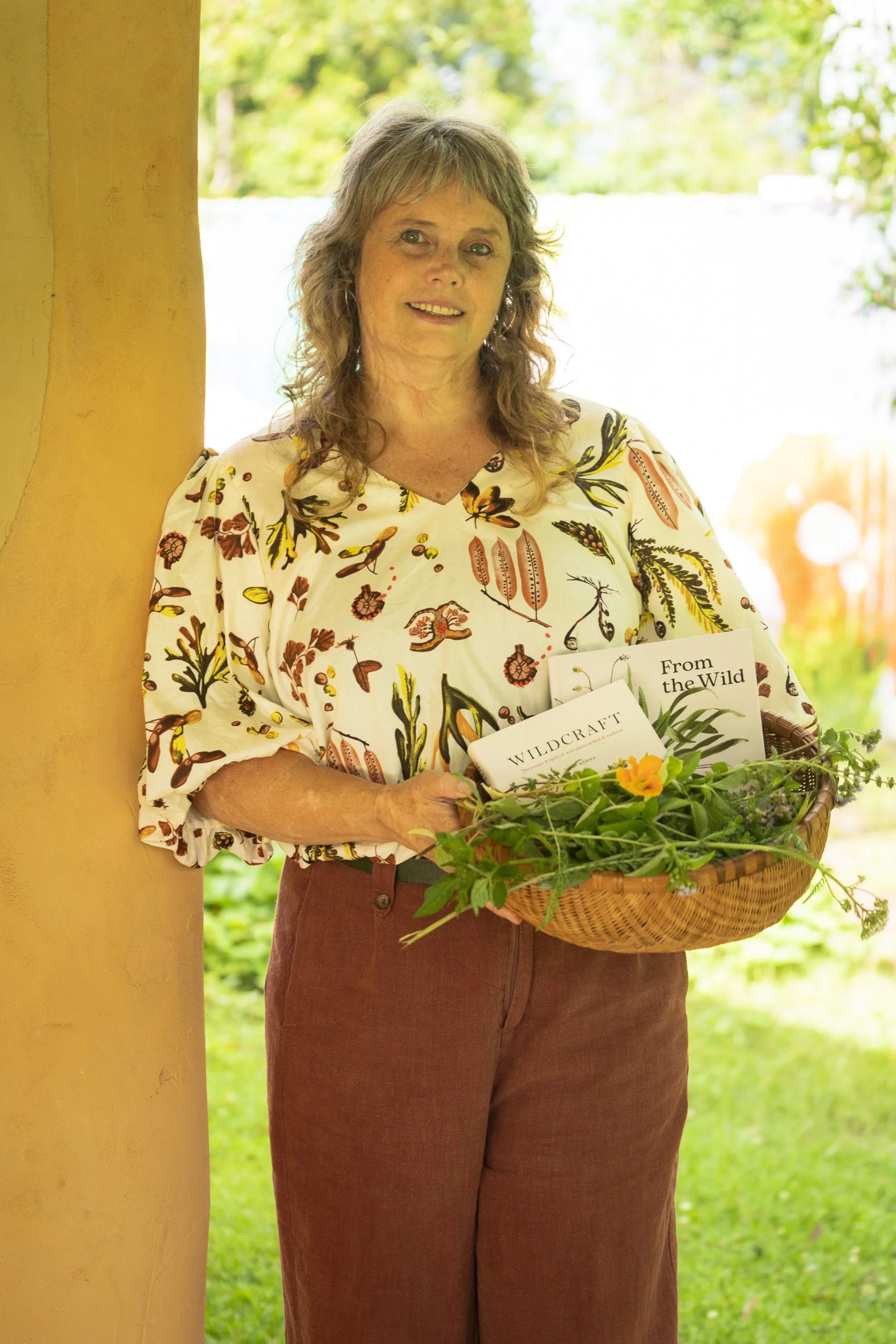A woman standing outdoors holding a basket of flowers and plants, wearing a floral blouse and maroon pants, near a yellow pillar with blurred green foliage in the background.