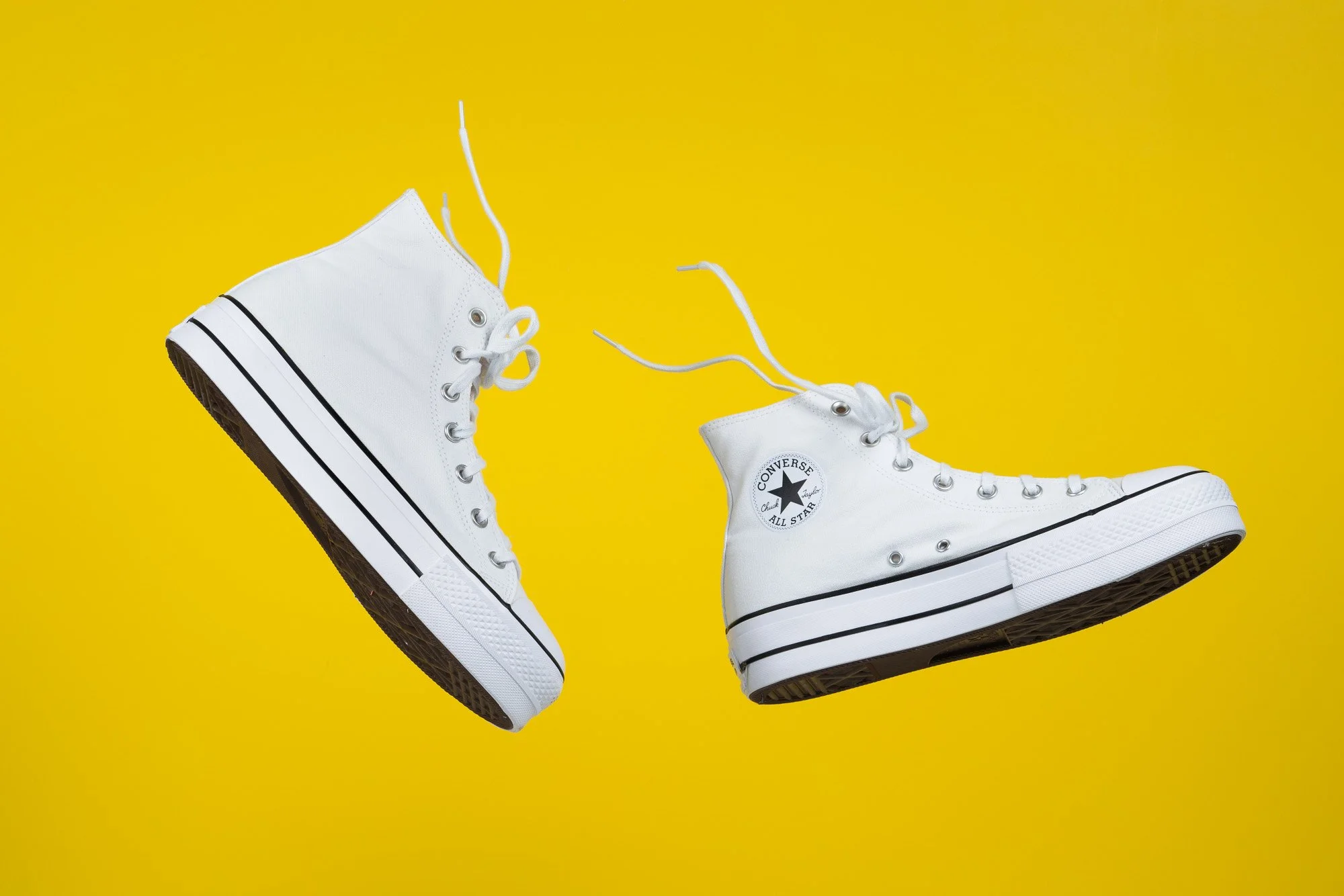 Pair of white high-top Converse sneakers with black stripes on the soles, set against a bright yellow background.