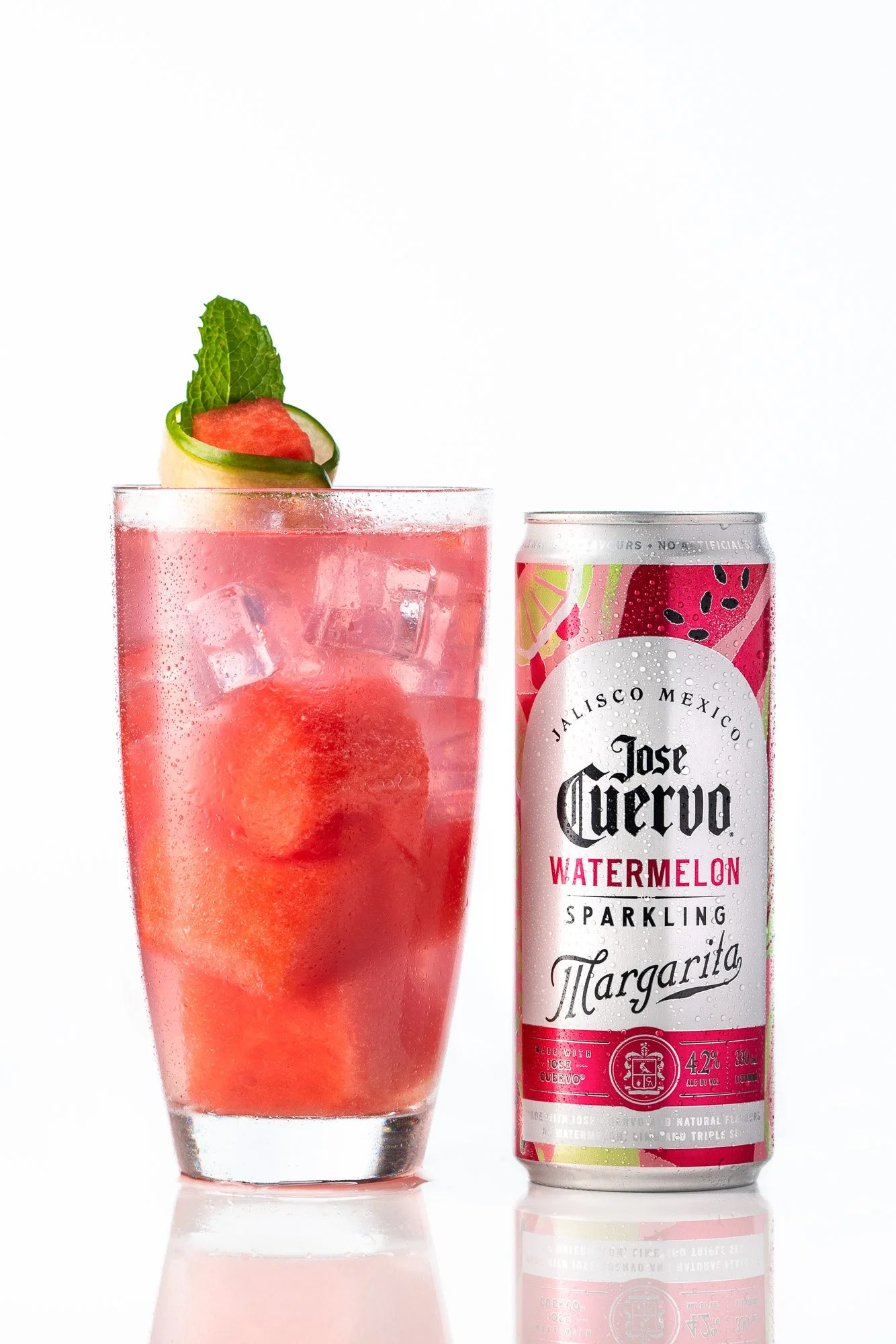 A tall glass filled with ice and a pink watermelon-flavored sparkling beverage, garnished with a slice of watermelon, a lime wedge, and a mint leaf; next to a can of Jose Cuervo Watermelon Margarita sparkling drink, with a colorful watermelon pattern