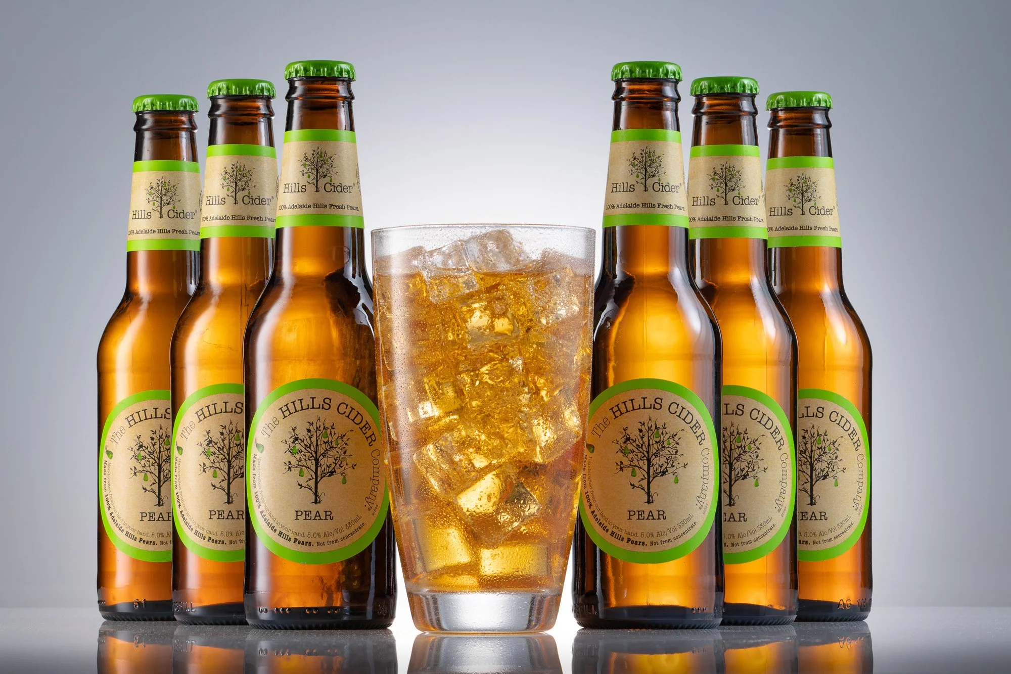 Six bottles of Hills Cider with a glass of cider and ice cubes in the middle, set against a plain background.