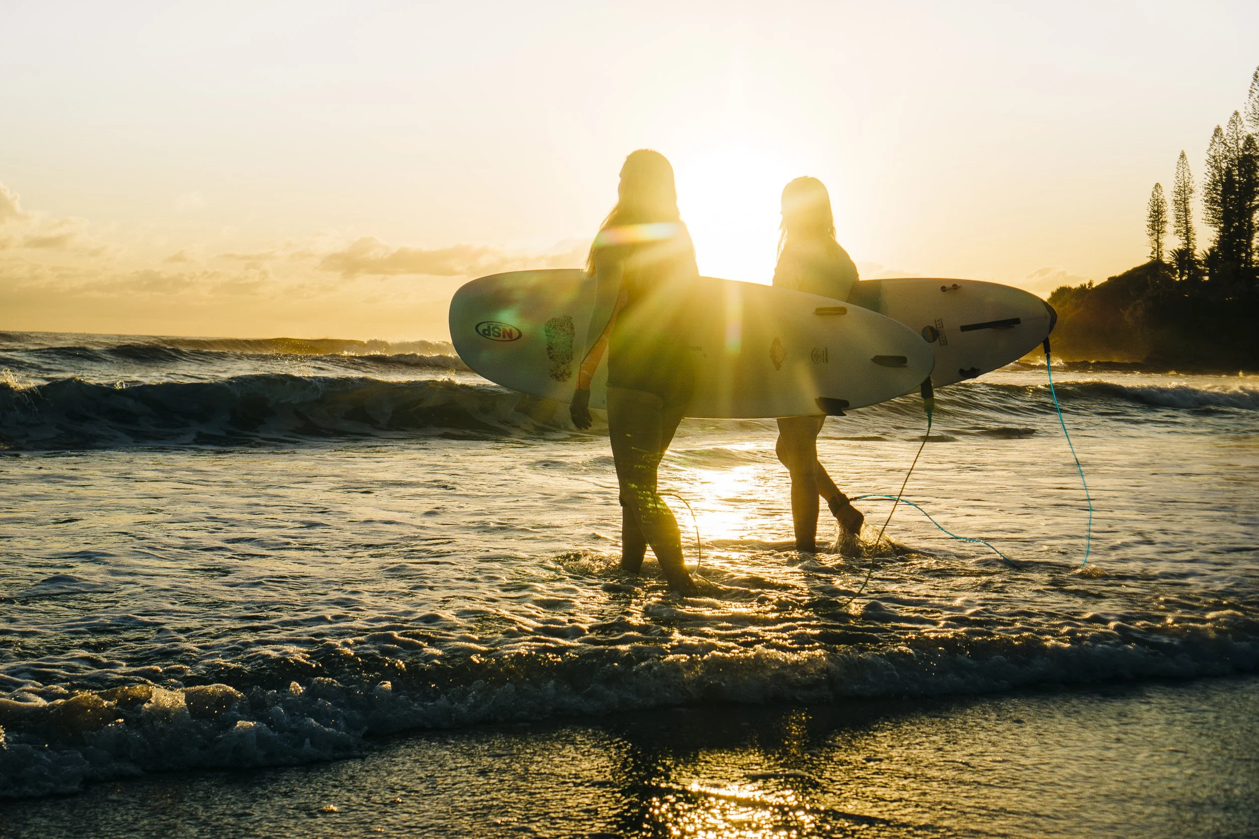 Two people carrying surfboards walking into the ocean at sunset, with the sun low on the horizon and silhouettes of trees on the shoreline.