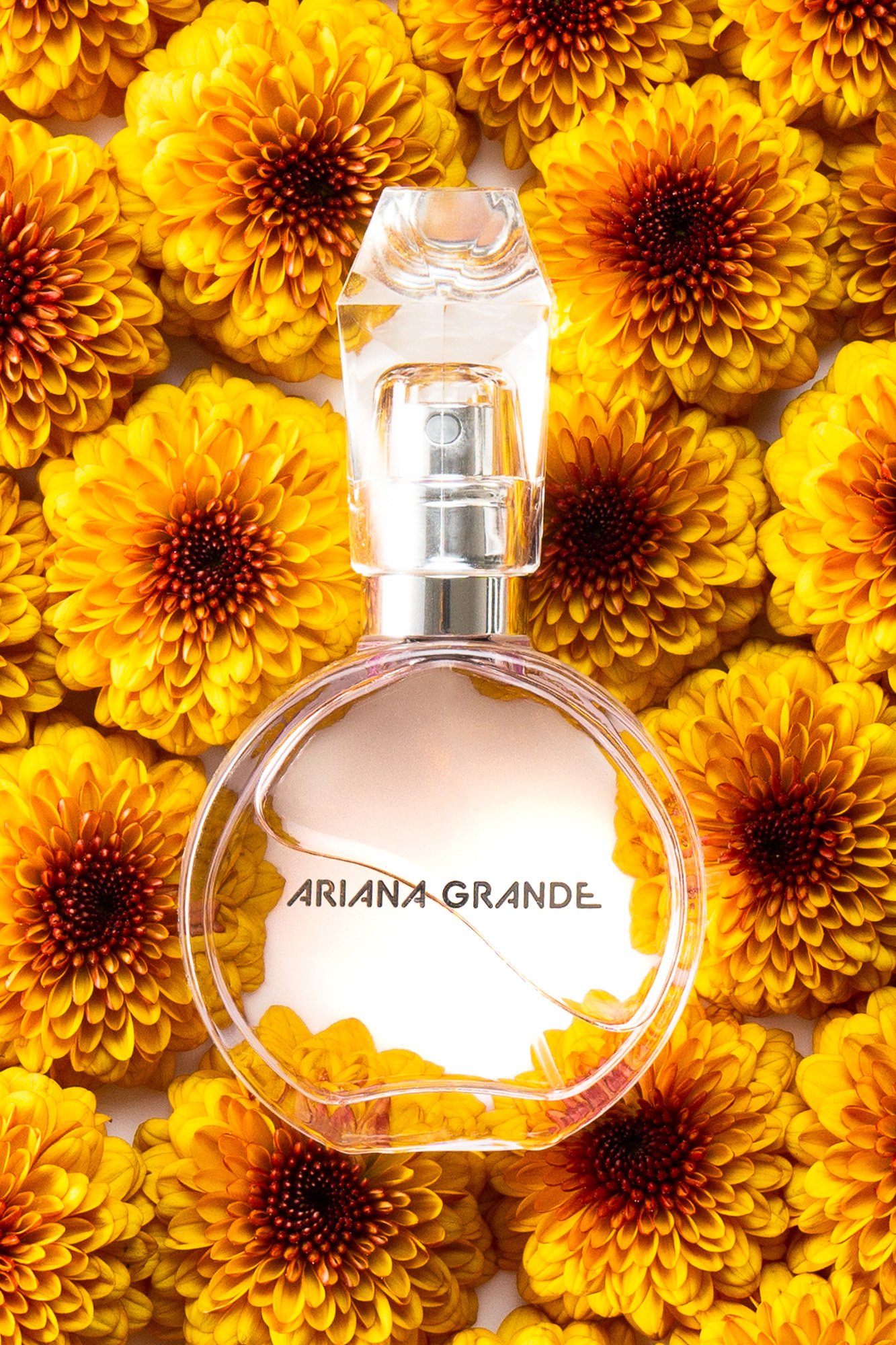 A perfume bottle labeled 'Ariana Grande' surrounded by yellow and orange chrysanthemum flowers.