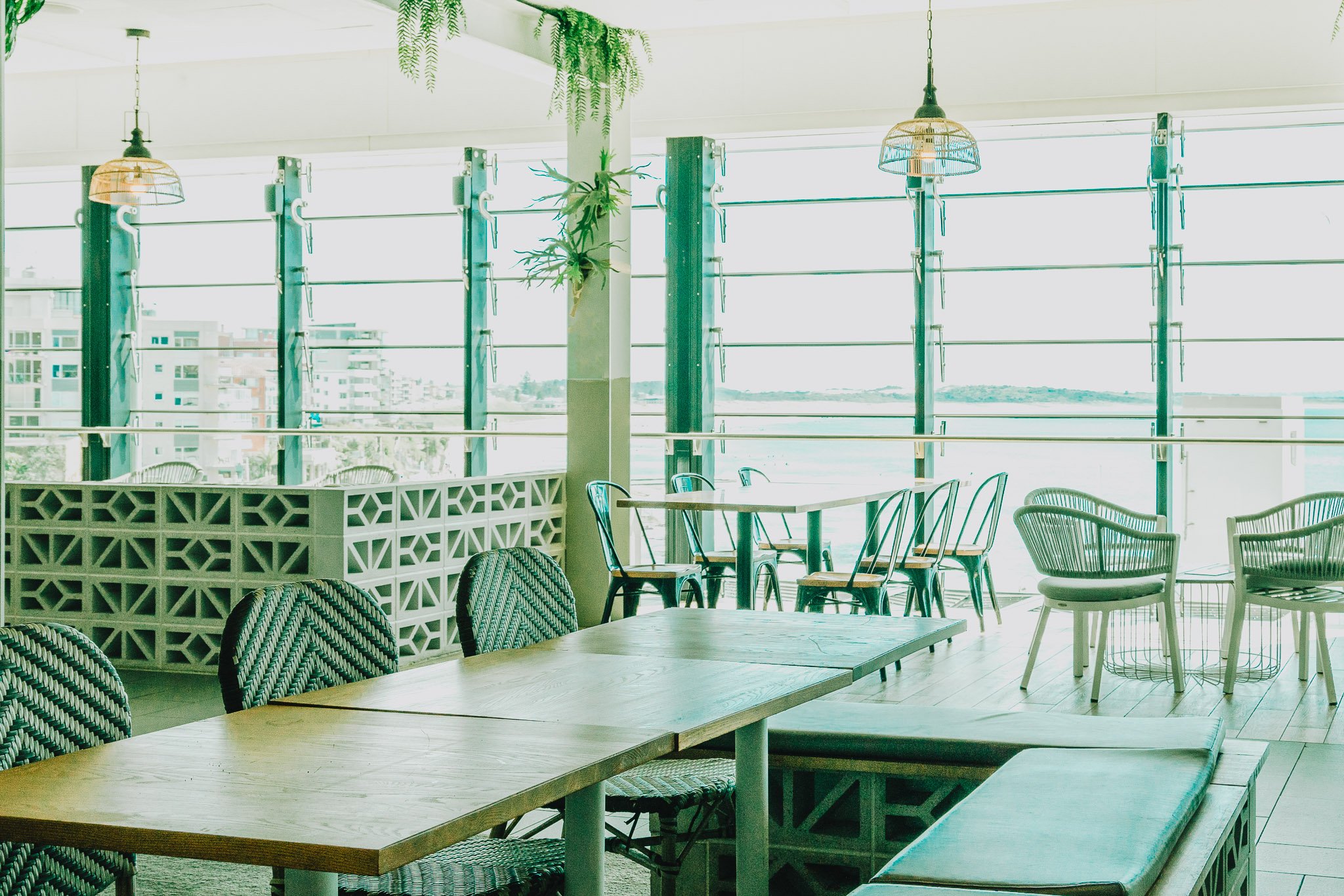 Interior of a modern cafe or restaurant with large windows, wicker chairs, wooden tables, hanging lamps, and greenery.