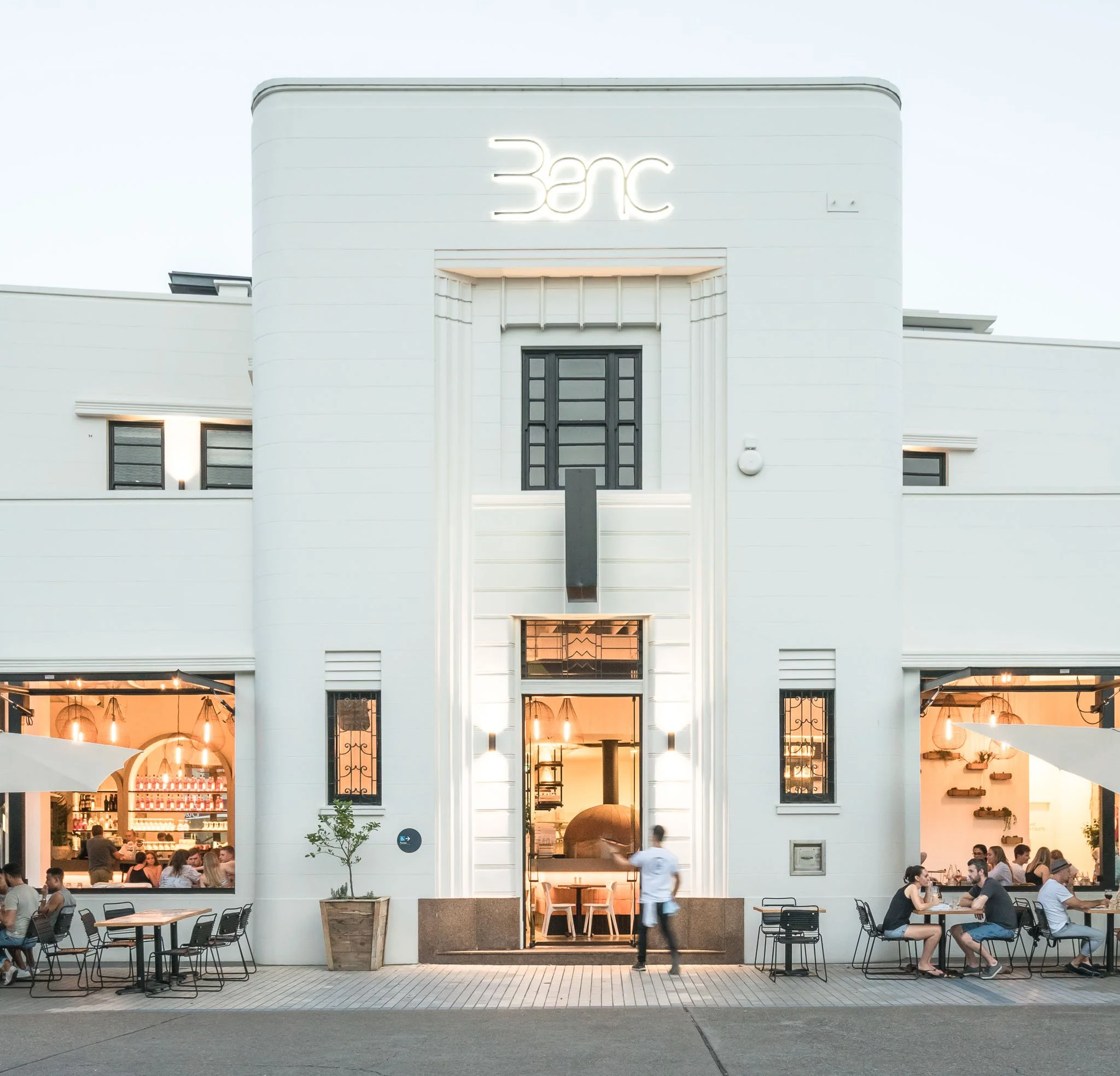 Modern white building with large glass windows, people dining outside at tables, and a neon sign above the entrance.