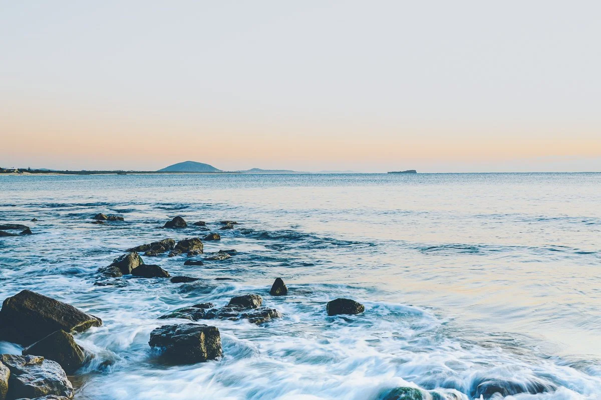 A calm ocean scene with rocks in the foreground and distant hills on the horizon at sunset.