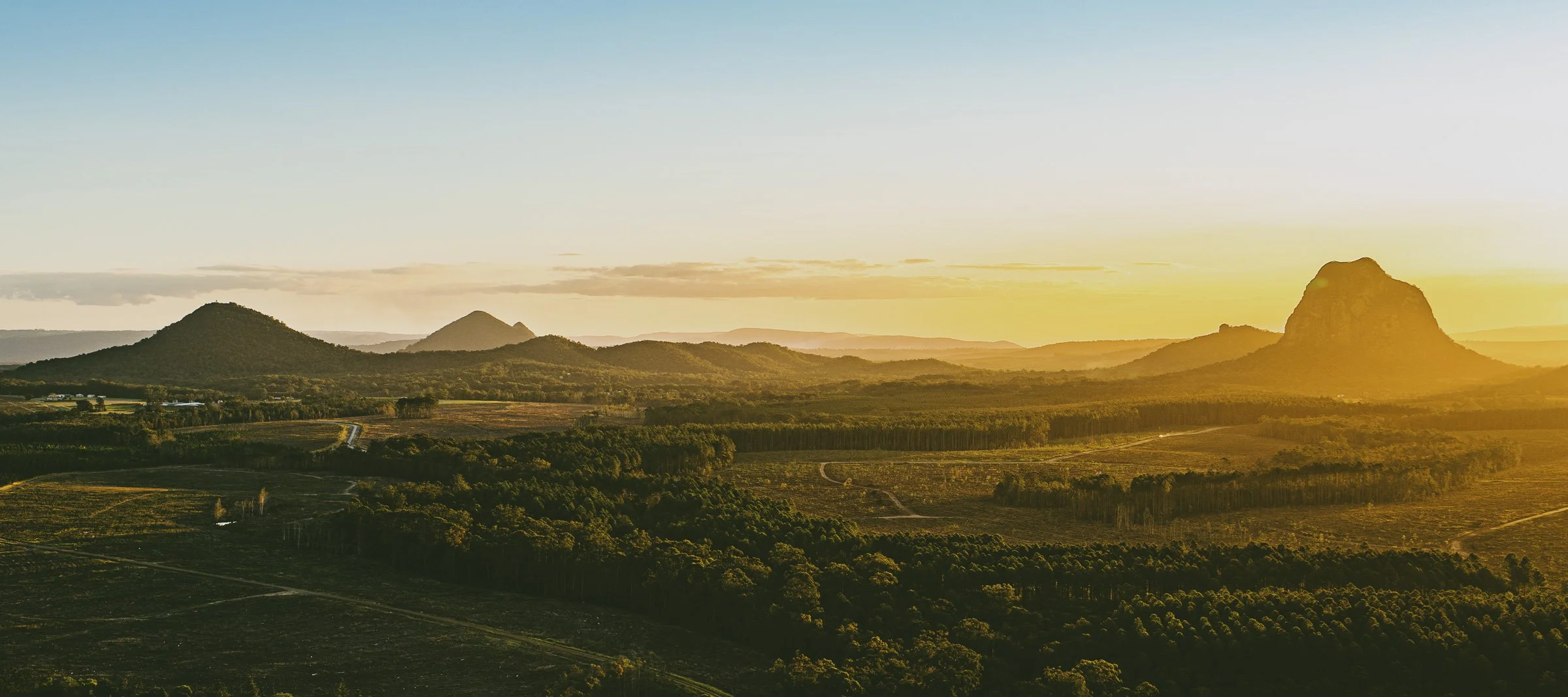 A landscape scene with several mountains or large hills at sunset, with a golden sky and a vast forested valley in the foreground.