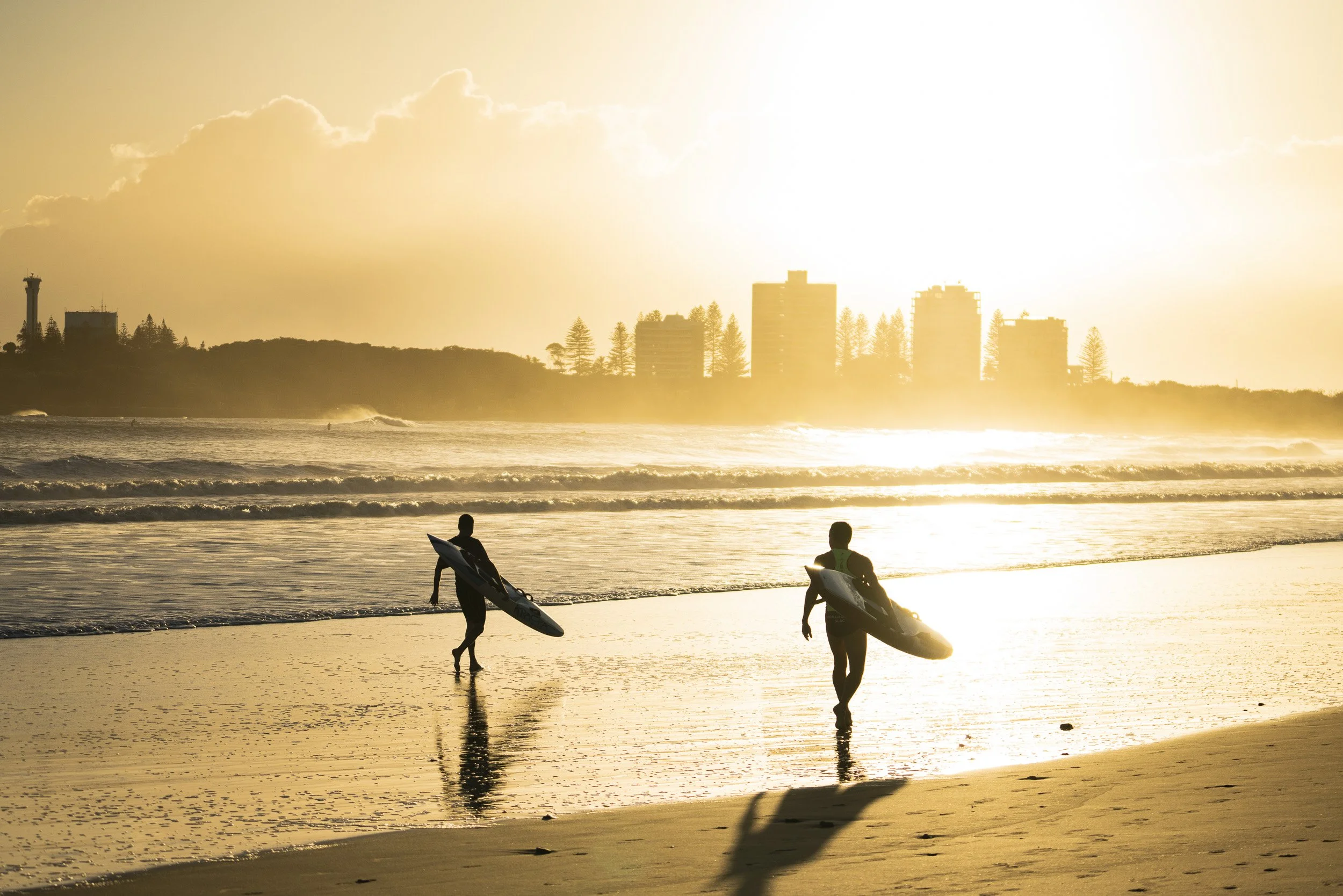 Silhouettes of two surfers walking along the beach at sunset, carrying surfboards with a city skyline and trees in the background, bathed in warm golden light.