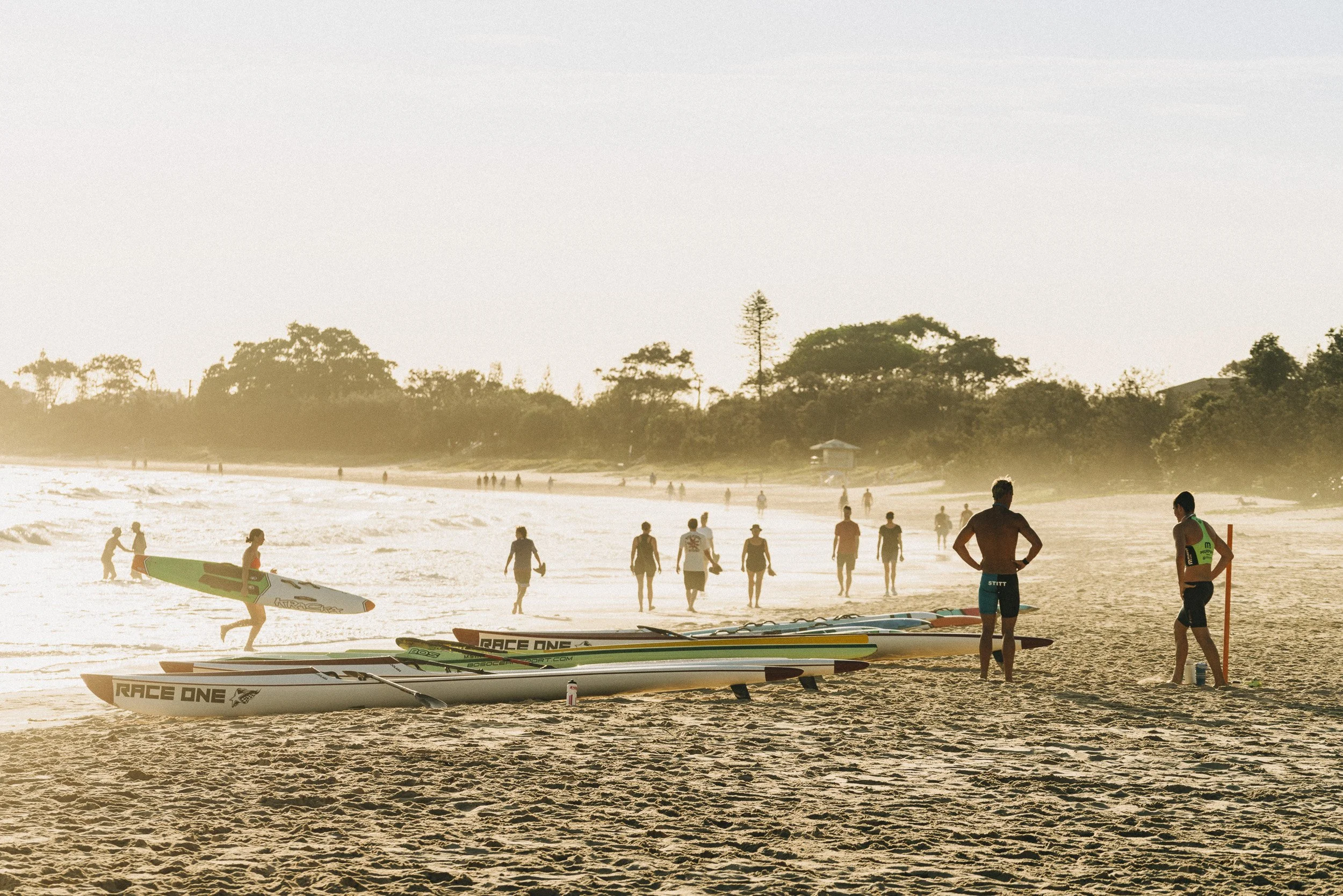 People at the beach preparing for surfing, with surfboards in the sand and others walking by the shoreline during sunset.