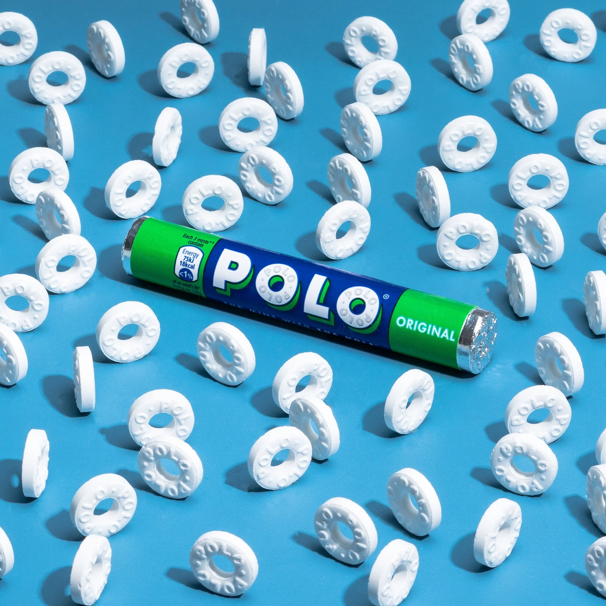A scattered collection of white Polo mints on a bright blue background, with a green and blue tube of Polo mints lying among them.