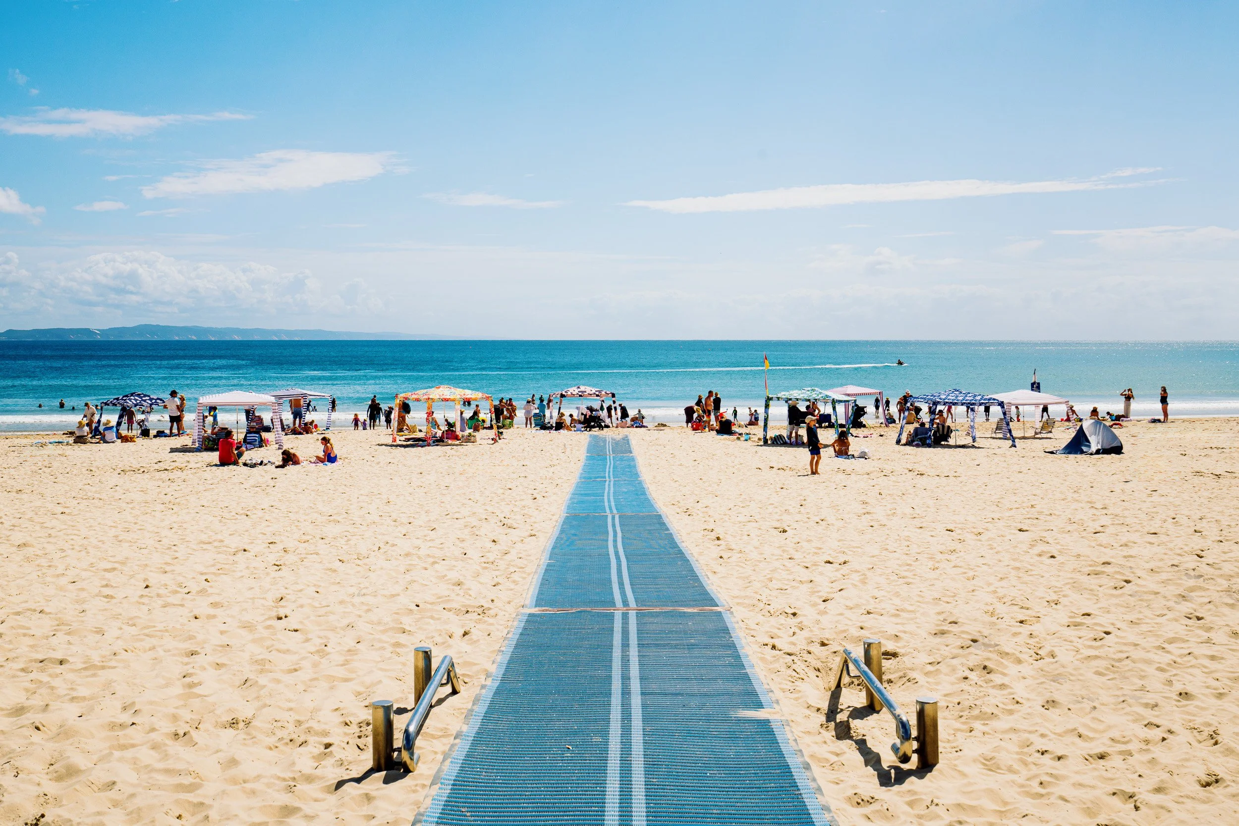 People at the beach under umbrellas, with a walkway leading toward the ocean, blue sky with some clouds, and a boat in the water.