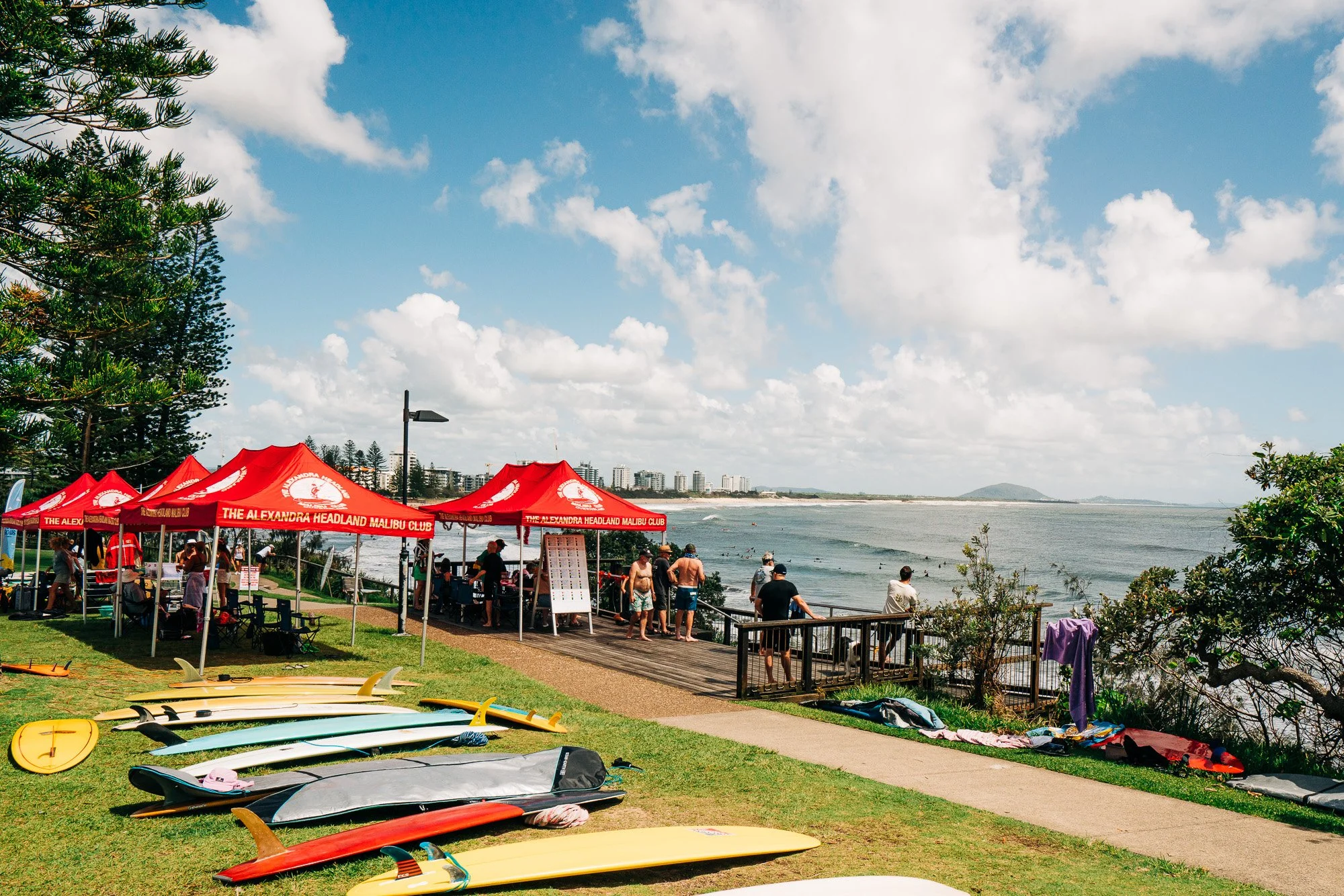 Beachside scene with red tents labeled 'The Alexandra Headland Malibu Club,' surfboards on the grass, people in swimwear near the water, and high-rise buildings in the background under a partly cloudy sky.