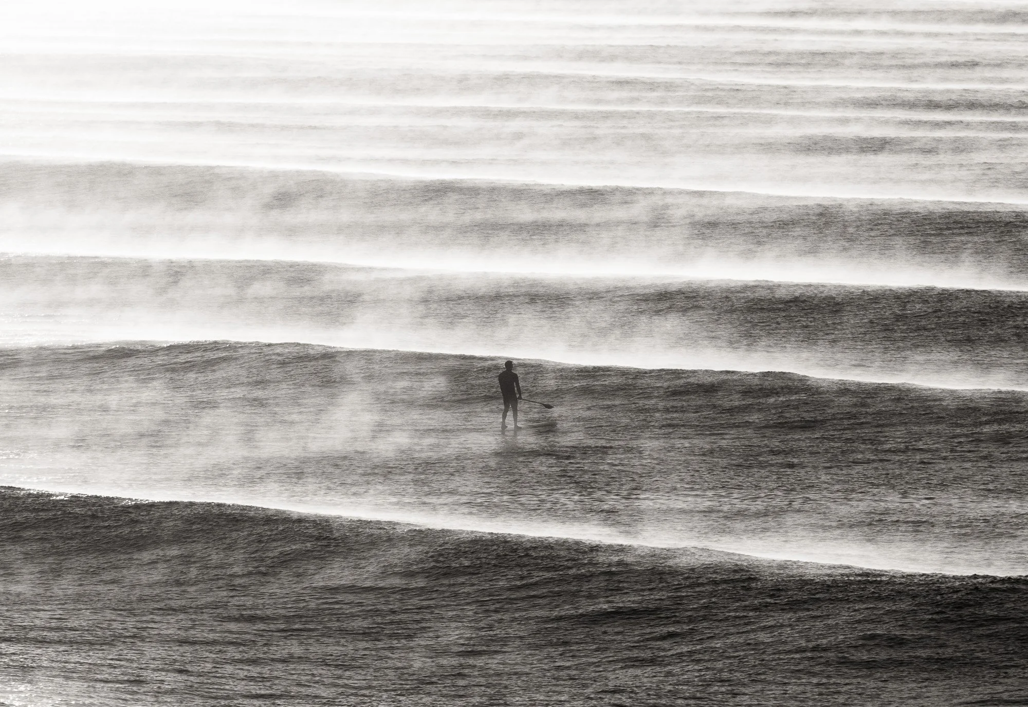 A person standing in the shallow surf of the ocean at sunset, with waves rolling in and mist rising from the water in a black and white photograph.