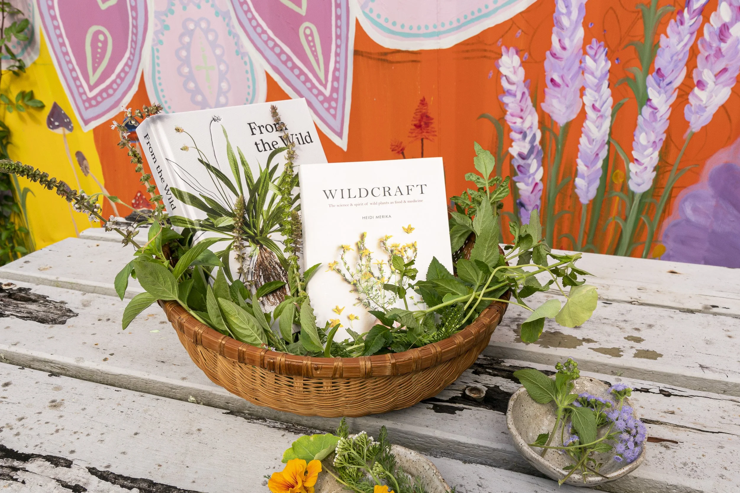 A woven basket filled with green herbs and flowers placed on a white distressed wooden table, with colorful painted backgrounds and smaller containers of flowers around it.