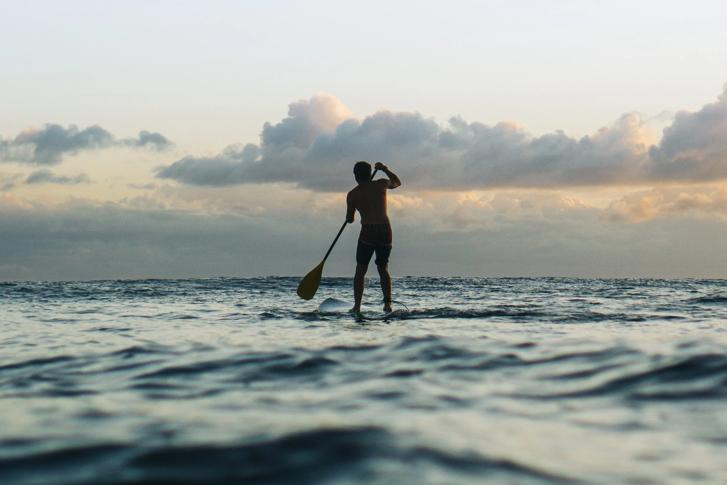 A person paddleboarding on the ocean at sunset with clouds in the sky.