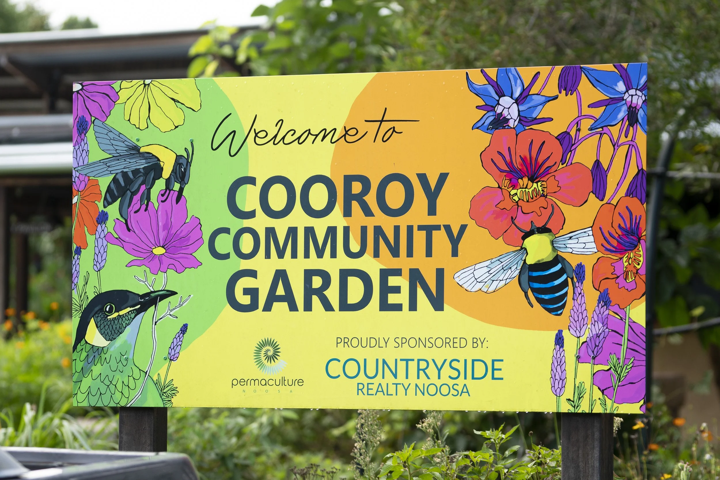 Colorful garden sign welcoming visitors to Coory Community Garden, decorated with illustrations of bees, flowers, and a bird.