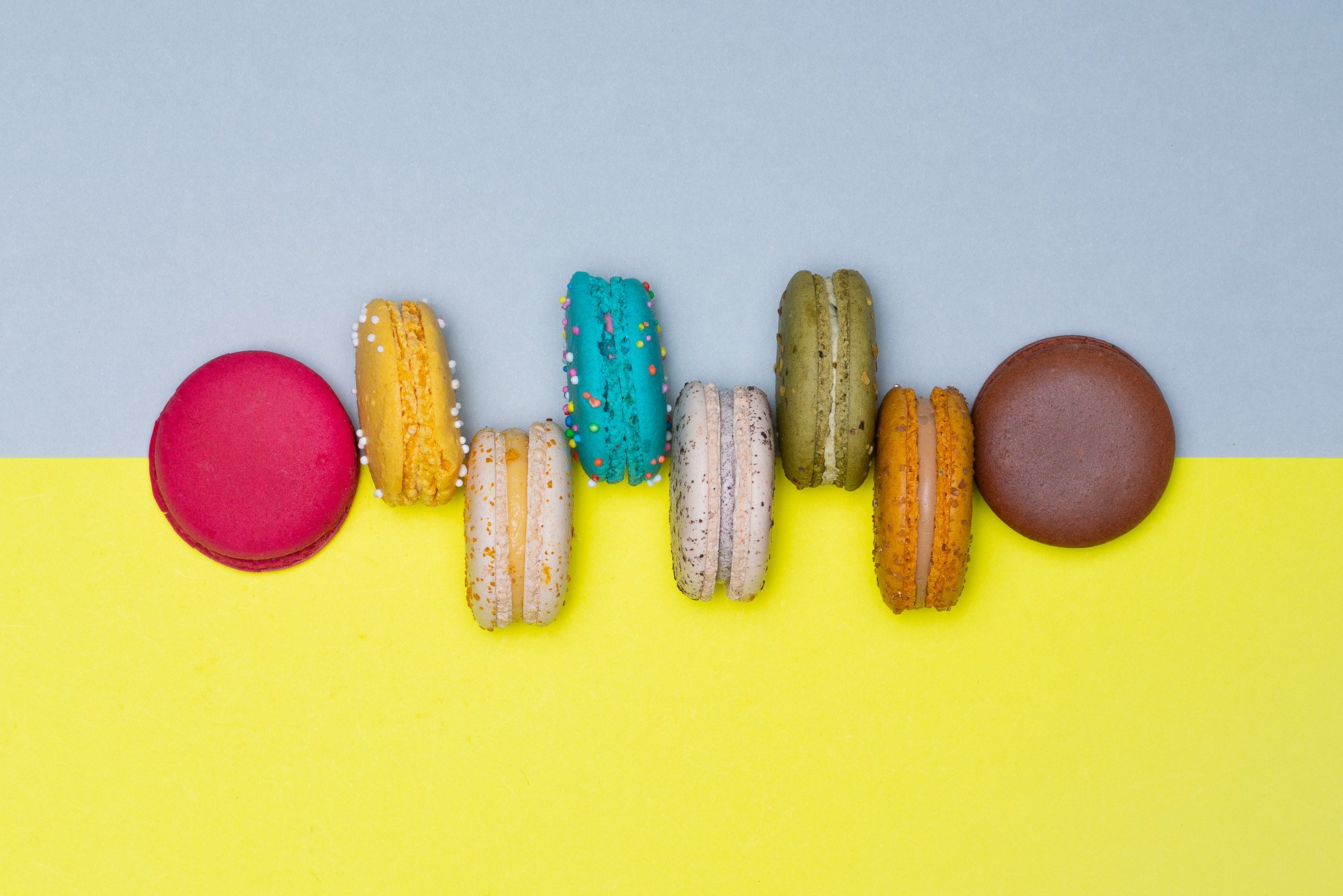 Colorful macarons lined up on a yellow surface under a light blue background.