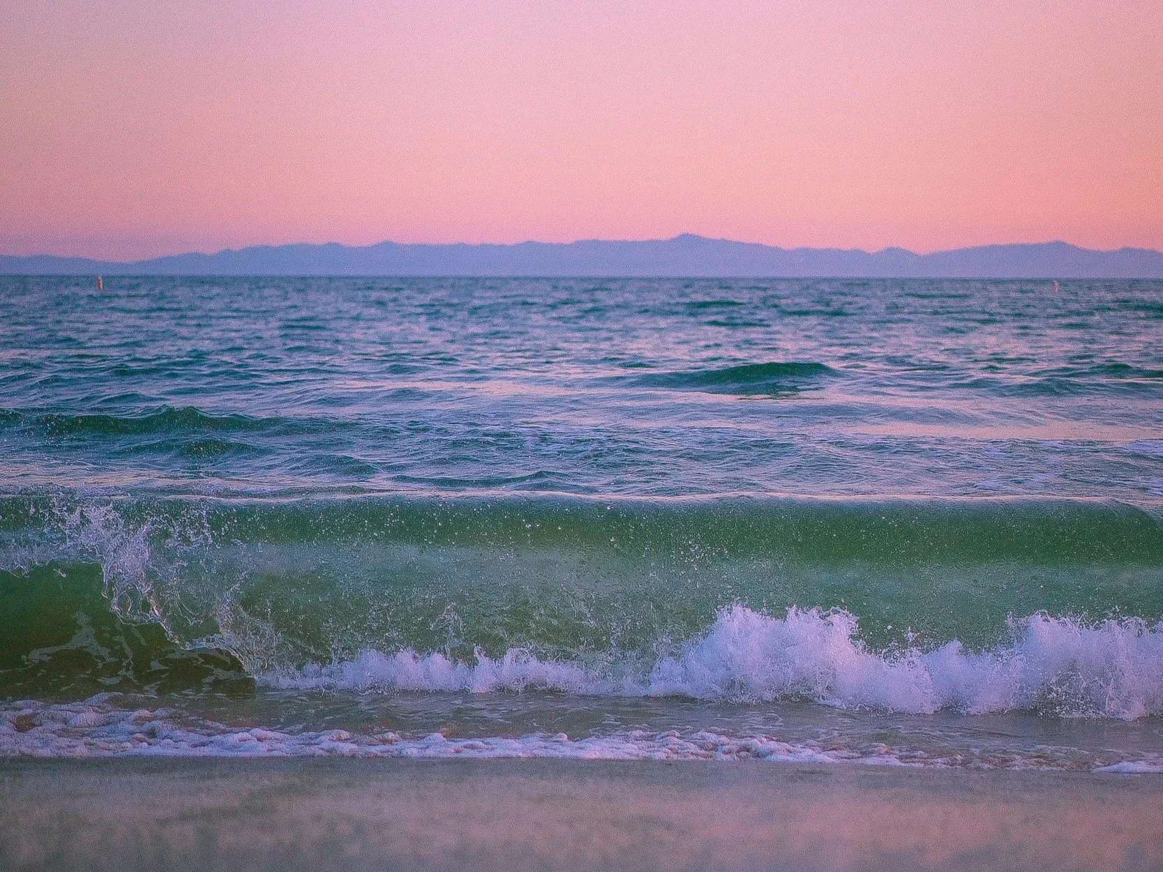 A serene view of the ocean at sunset with waves crashing onto the shore, pink and purple sky, and distant mountain silhouette on the horizon.