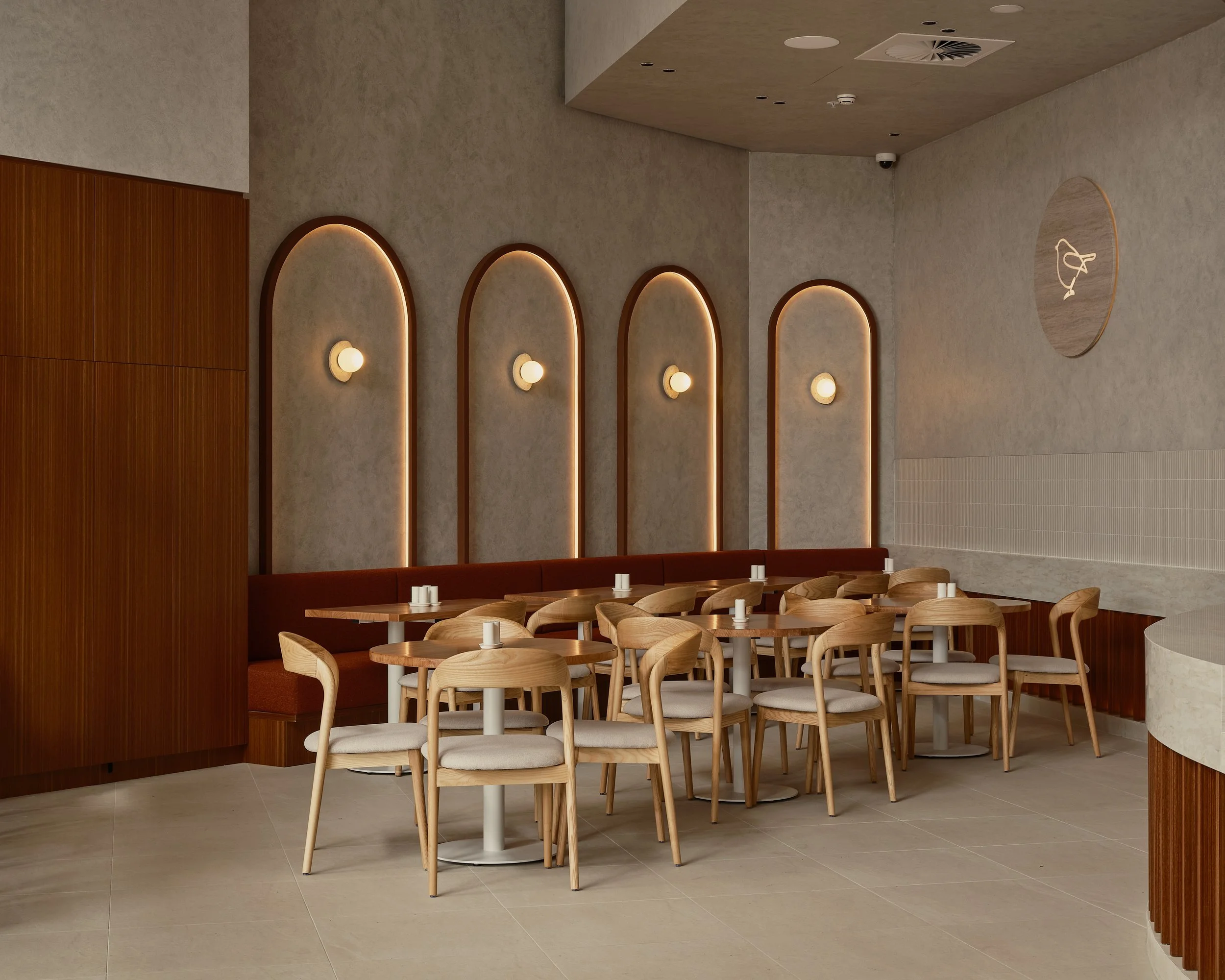 Empty dining area with round tables and wooden chairs, beige walls with arched wooden accents, wall-mounted lights, and a textured ceiling with a ceiling vent.