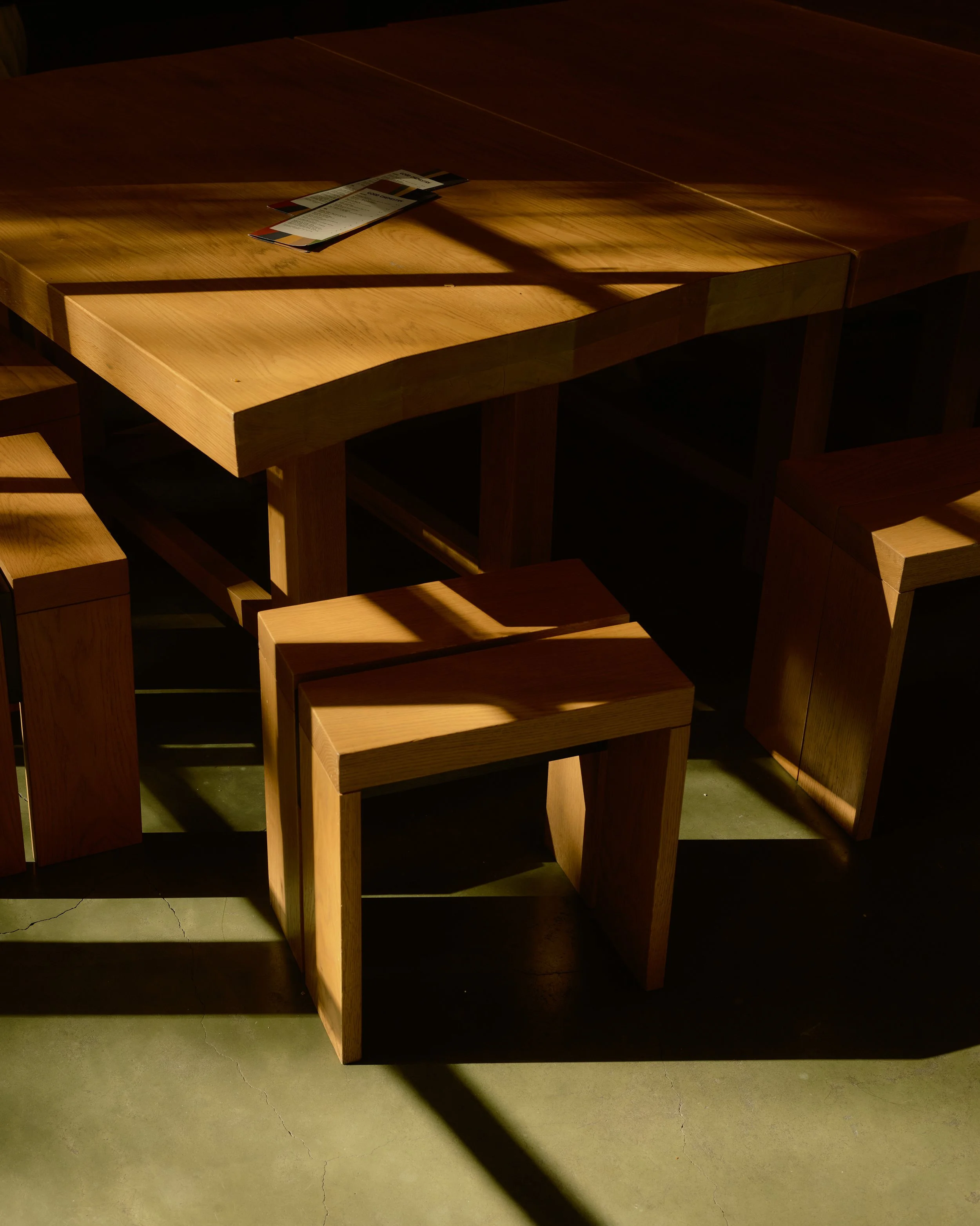 A wooden dining table with a natural finish, with a few paper receipts or menus placed on top. The table is located in a dimly lit space, with shadows cast across the surface and the floor.