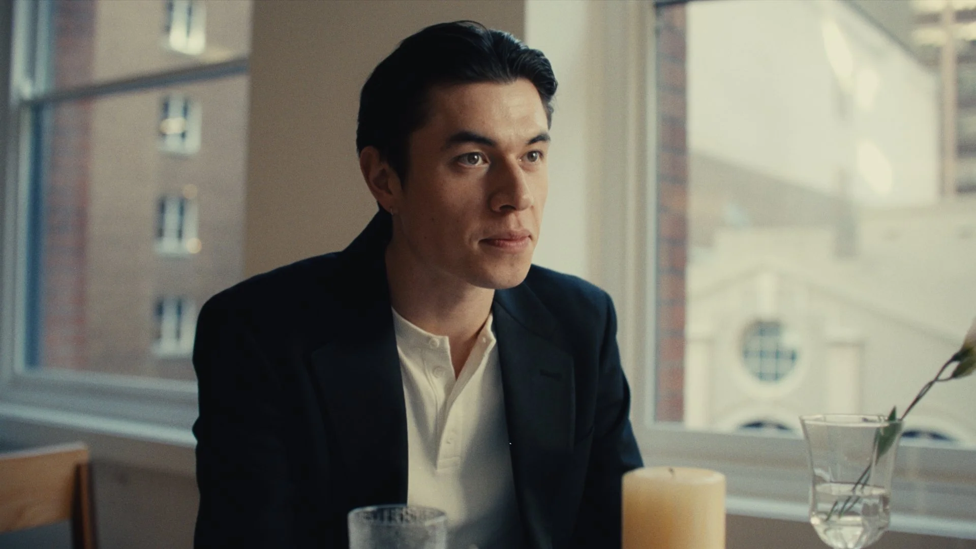 A man with dark hair wearing a black blazer and white shirt, sitting at a table near a window in a bright room.
