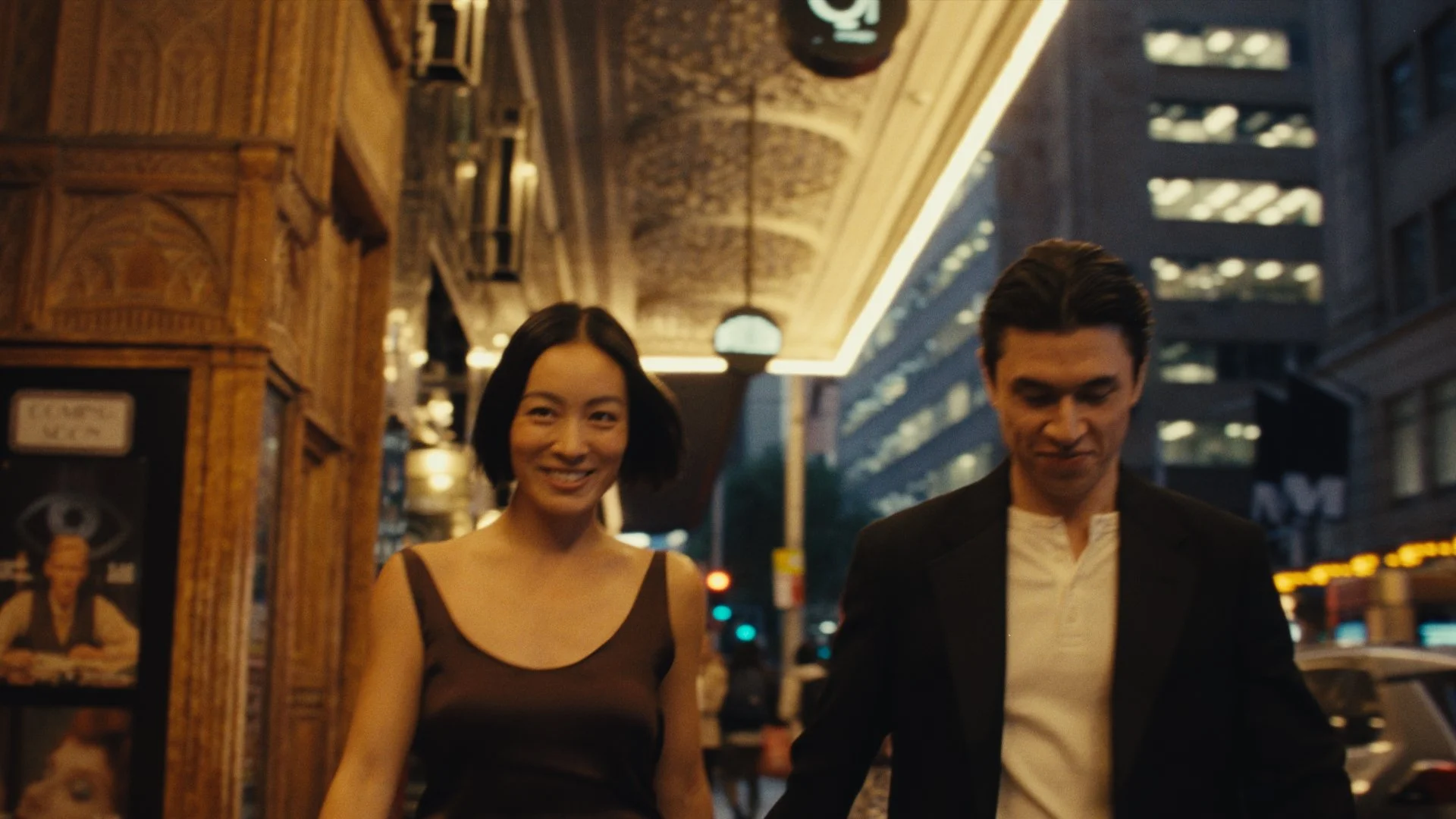 A woman in a brown dress with spaghetti straps and a man in a white shirt with a black blazer walking together on a city street at night, lit by streetlights and store signs.
