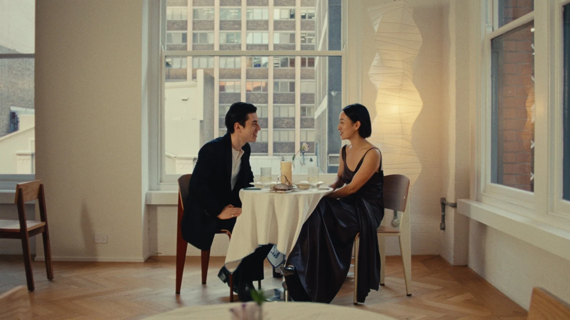 A man and woman are sitting at a small, round table in a bright room with large windows. They are smiling at each other, and the table is set for two with drinks and a small flower. The background shows an urban cityscape. The woman is wearing a black dress, and the man is in a black suit.