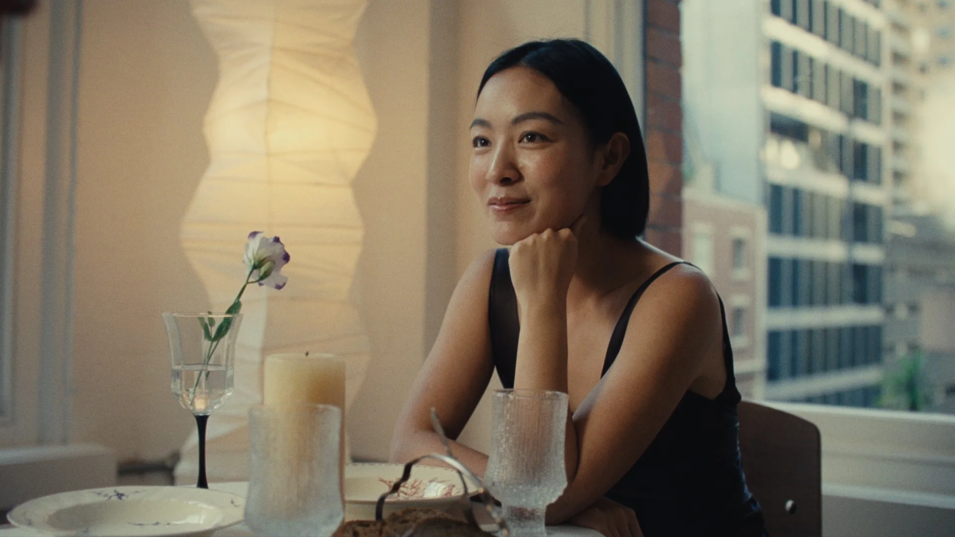 A woman sitting at a dining table with a window behind her, smiling gently. The table has various items, including a glass of water, a white plate, a vase with a purple flower, and candles. She has short black hair and is wearing a black sleeveless top.