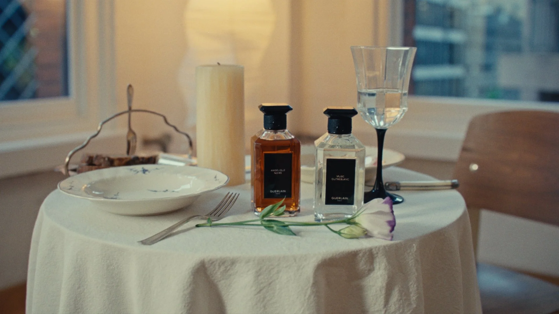 A round table covered with a white tablecloth holds two perfume bottles, an empty wine glass, a fork, a flowering branch, a white plate, and a candle. The background includes a window with a blurry city view and a lit lamp.