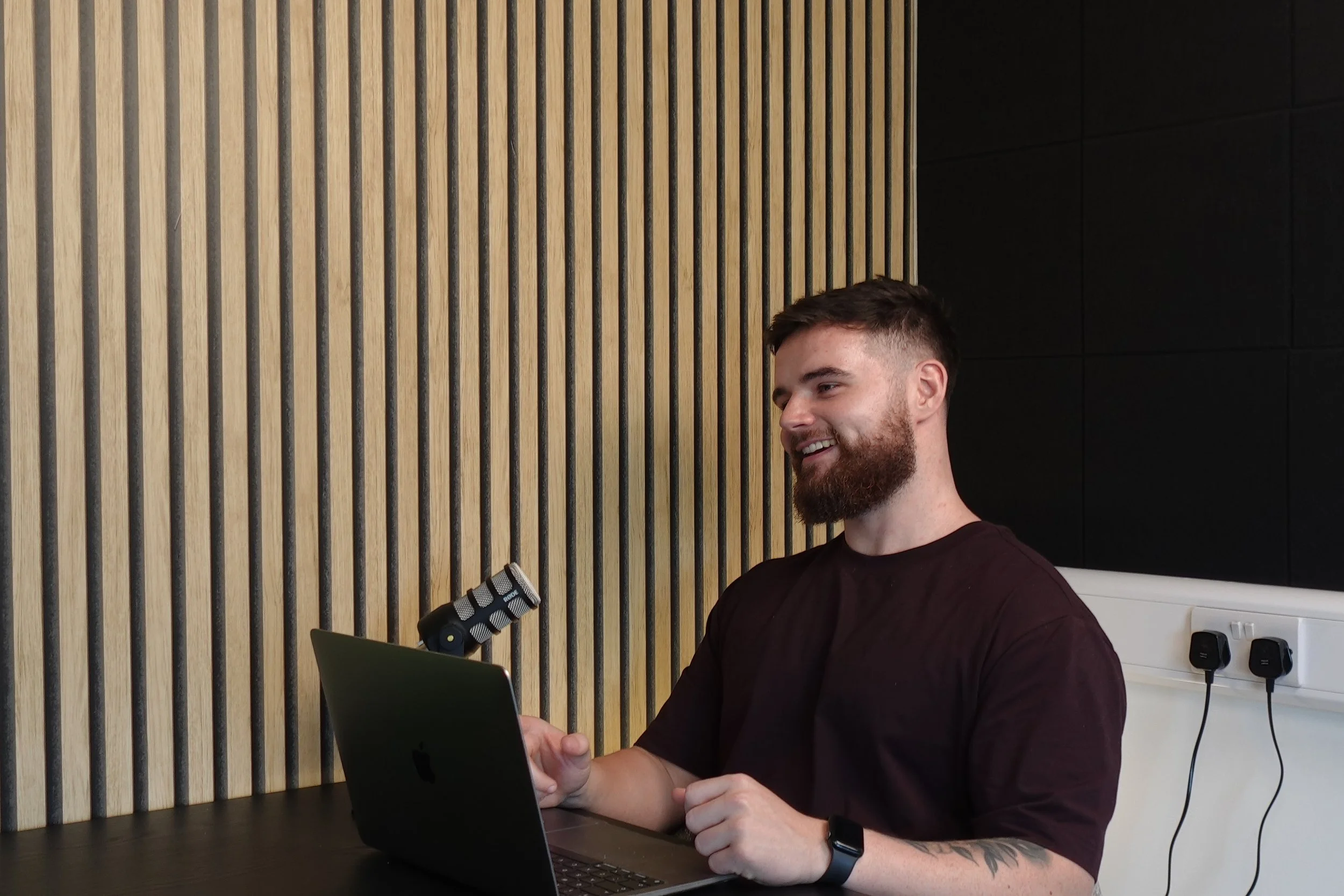A man with a beard and short hair, wearing a black shirt and smartwatch, sitting at a black table in front of a laptop. He is smiling and shaking hands with someone out of the frame. The background features a wooden slat wall and a black wall with electrical outlets and a power strip.