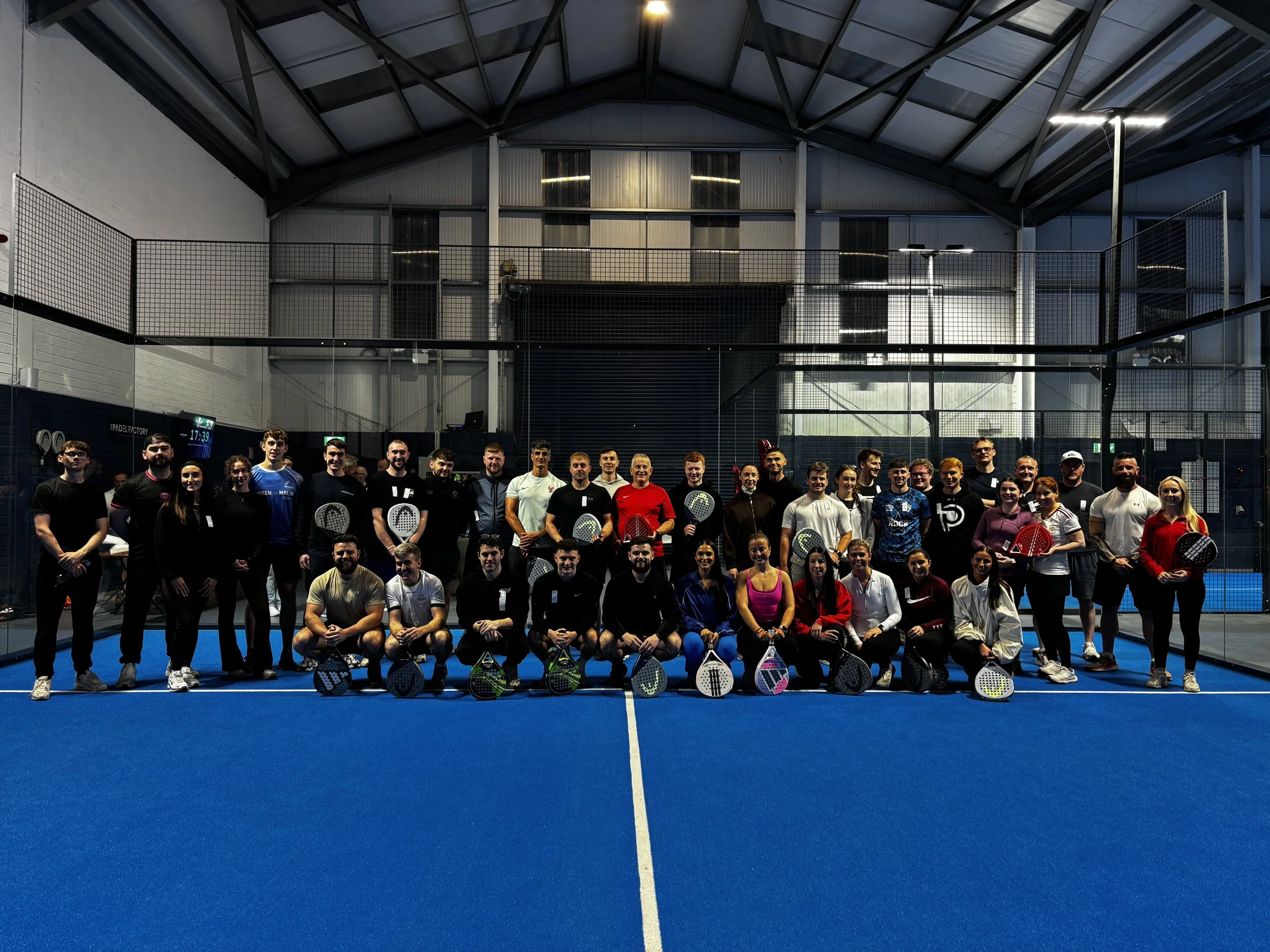 Group of people standing on a blue indoor padel tennis court, holding paddles, smiling for a group photo, with a high ceiling and windows in the background.