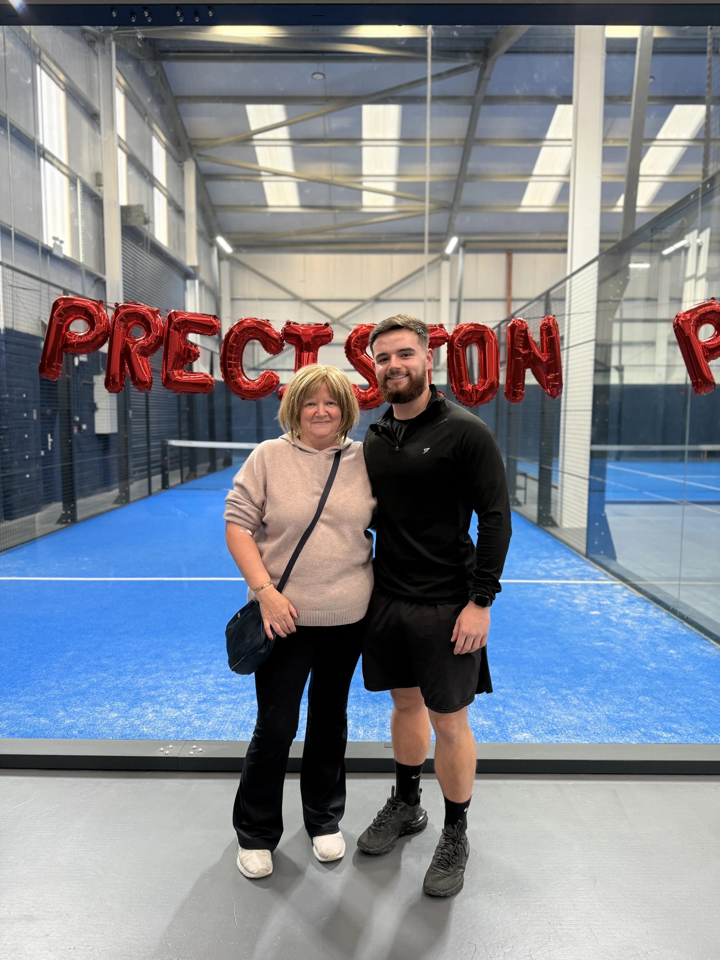 A woman and a man standing together on a blue tennis court indoors. They are smiling, and the woman has blonde hair and is wearing a beige hoodie and black pants. The man has a beard, short hair, and is wearing a black athletic jacket and black shorts. In the background, red balloons spell out the word 'PRESOLUTION'.