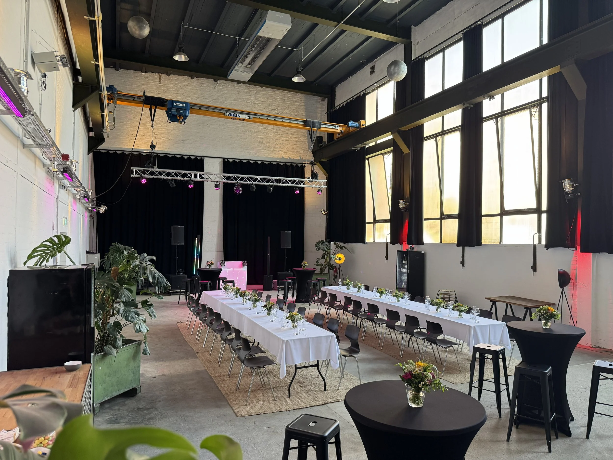 Der Saal ist für eine Veranstaltung vorbereitet, mit langen Tischen, die mit weißen Tischdecken bedeckt sind und kleine Blumenarrangements haben. Es gibt schwarze Stühle um die Tische, Pflanzen in der Ecke, eine Bühne mit einer schwarzen Kulisse und Monitoren, sowie eine Deckenkran für Technik. Große Fenster lassen natürliches Licht herein.