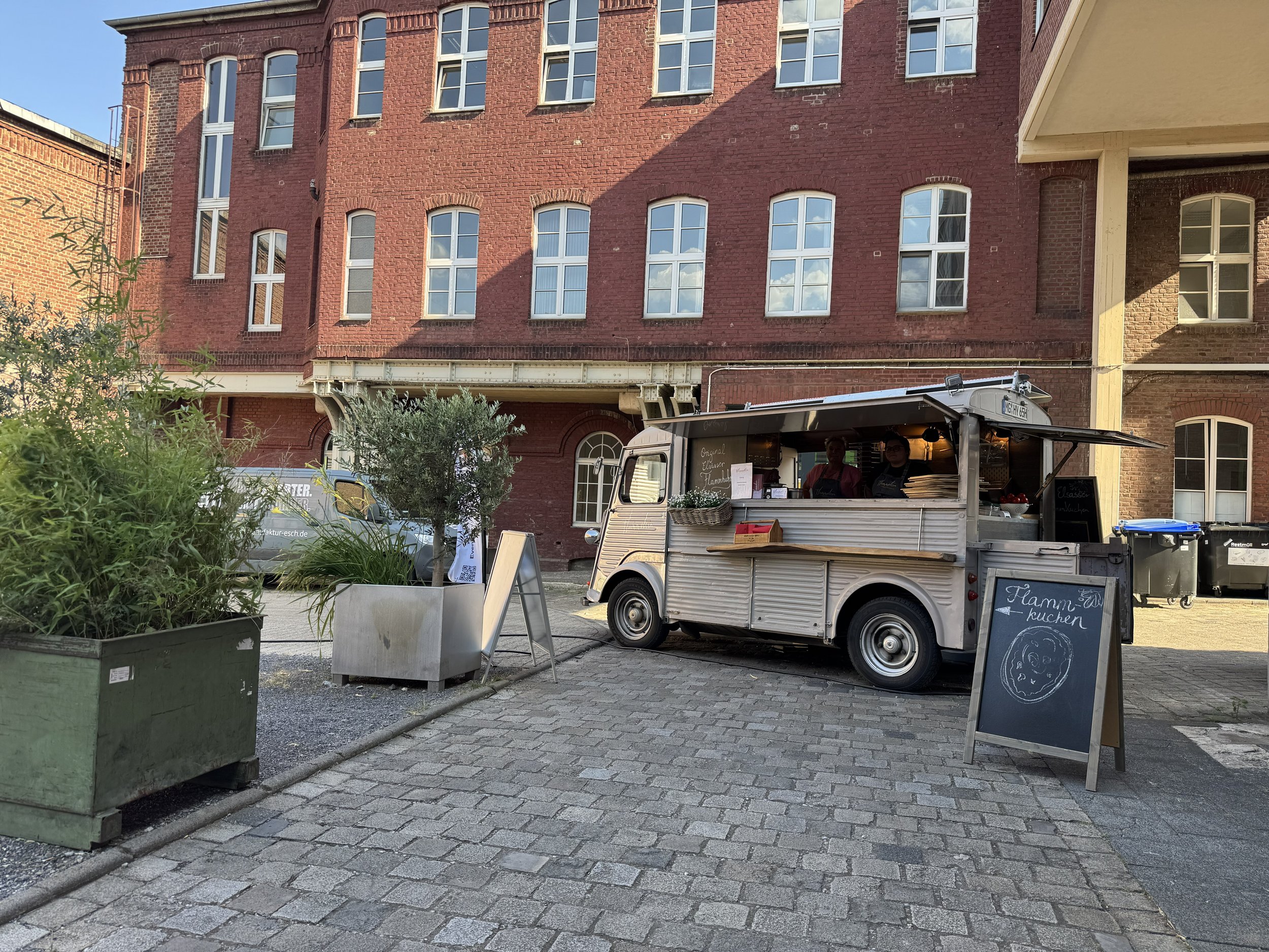Straßenverkaufswagen für Flammkuchen vor rotem Backsteingebäude, mit Beschilderung, Pflanzen in großen Töpfen, und Menschen im Hintergrund.