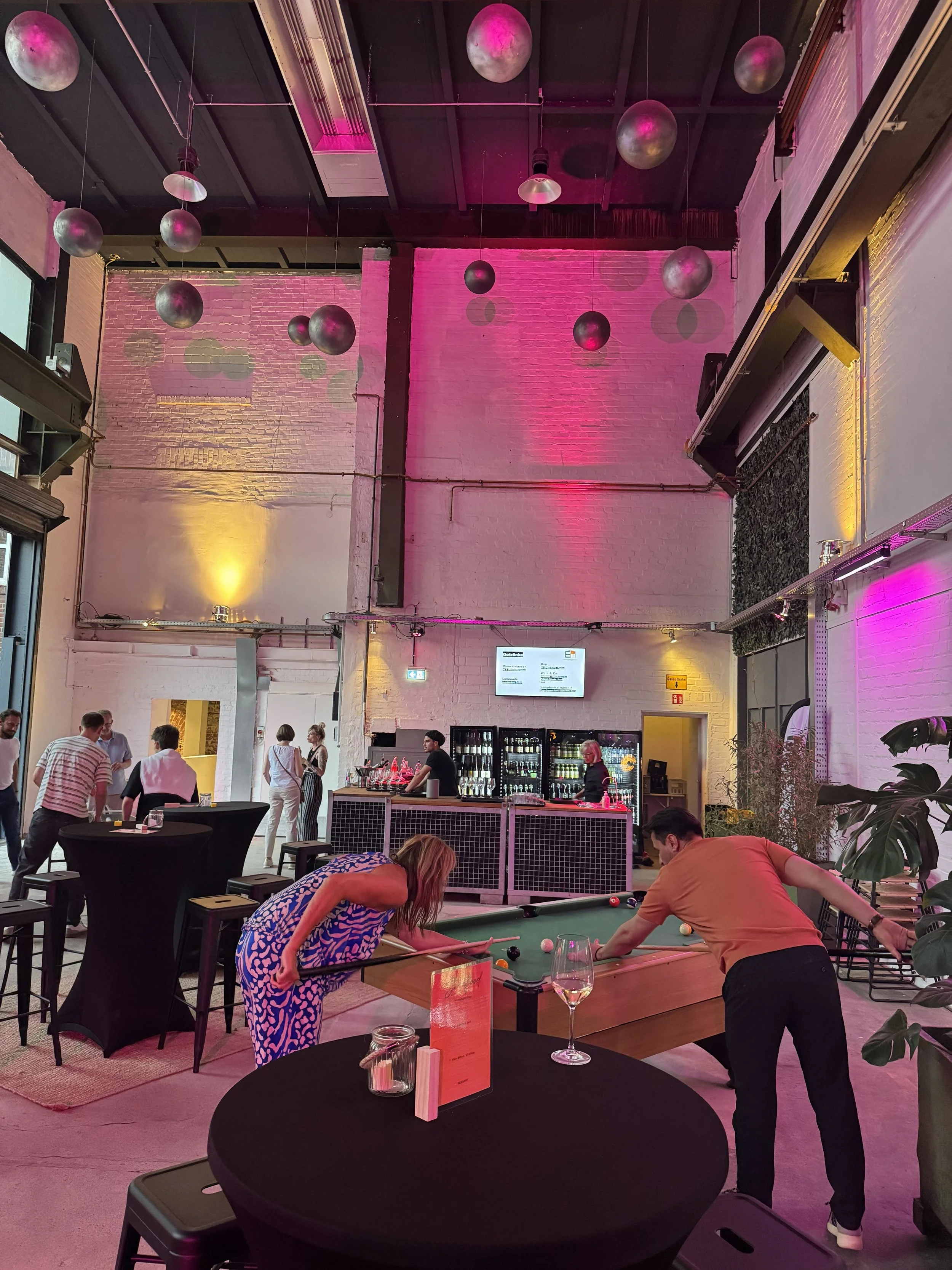 Innenaufnahme einer modernen Bar mit schwarzen und weißen Wänden, pinker Beleuchtung an der Decke und mehreren schwarzen runden Tischen. Es sind Menschen, die Billard spielen, und an der Bar stehen mehrere Gäste. Es gibt eine Bar mit Kühlschrank und Getränken.