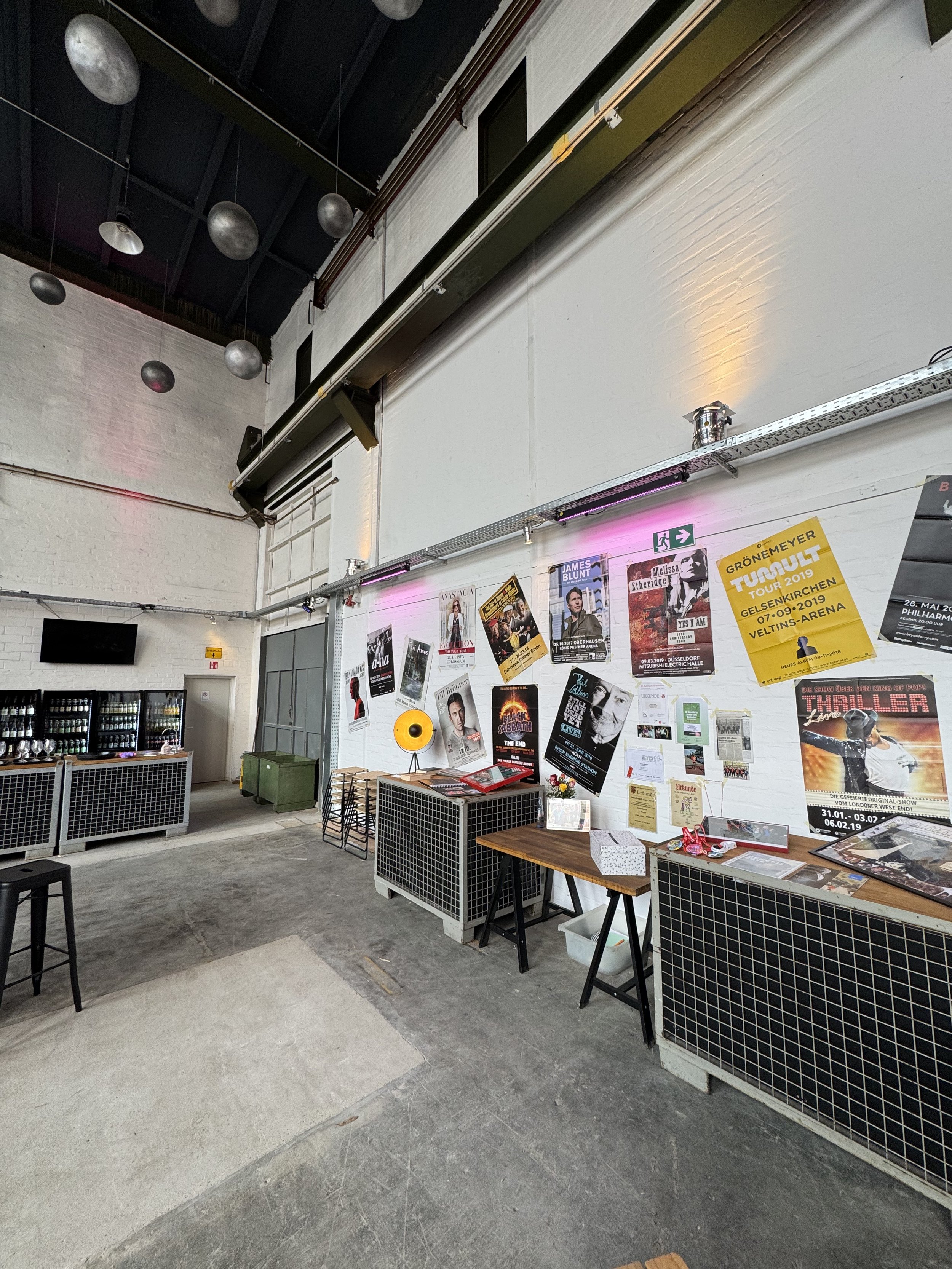 Innenansicht einer Veranstaltungshalle mit Plakaten an der Wand, einigen Tischen, Stühlen und Getränkekühlschränken, an der Decke hängen Dekorations-Objekte und es gibt eine Fluchtwegleitung an der Wand.