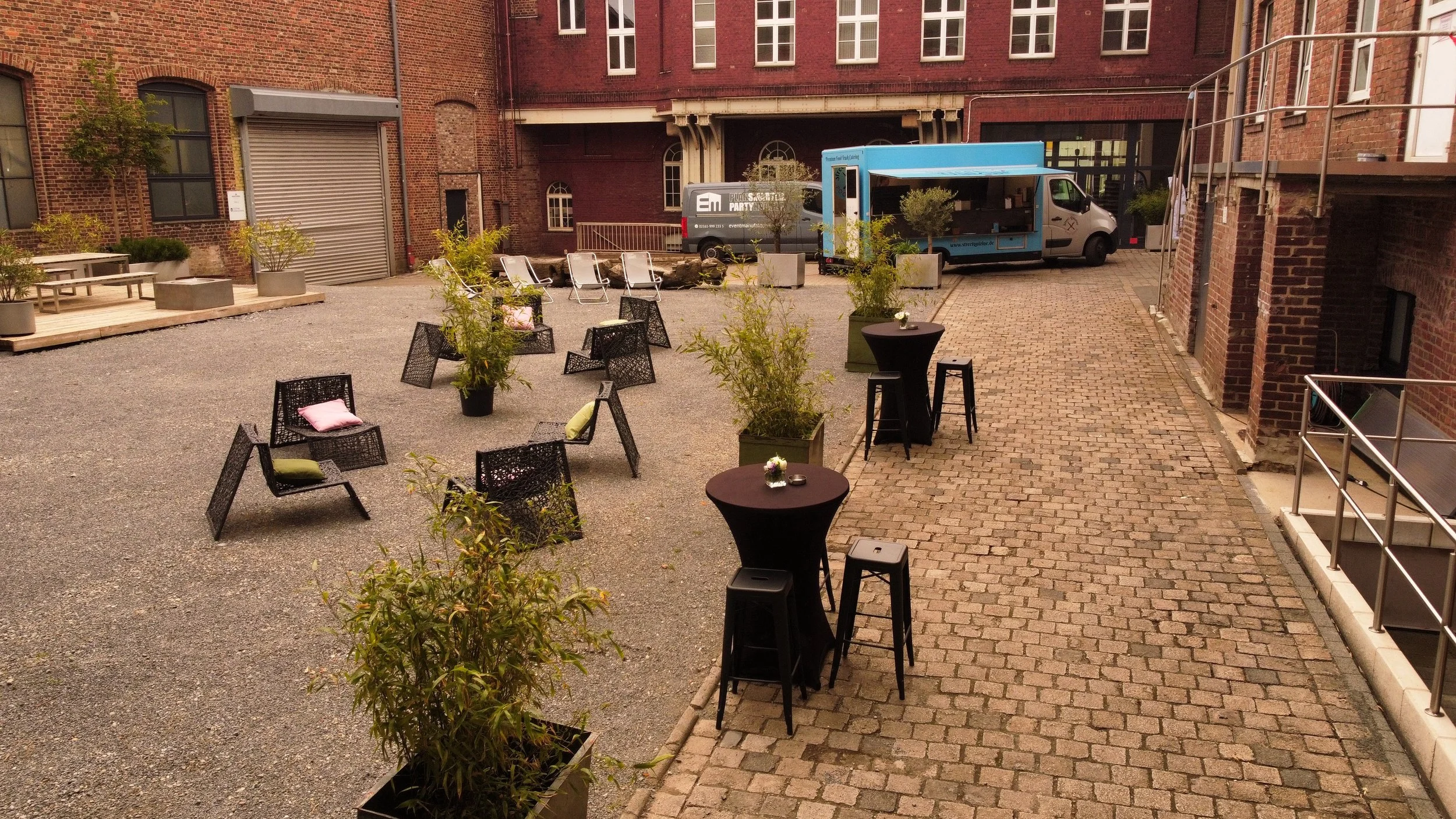 Ein Innenhof mit Sitzgelegenheiten, einigen Pflanzen in großen Töpfen, Holztischen und Stühlen, sowie einem Foodtruck und einem Lieferwagen vor roten Backsteinwänden.