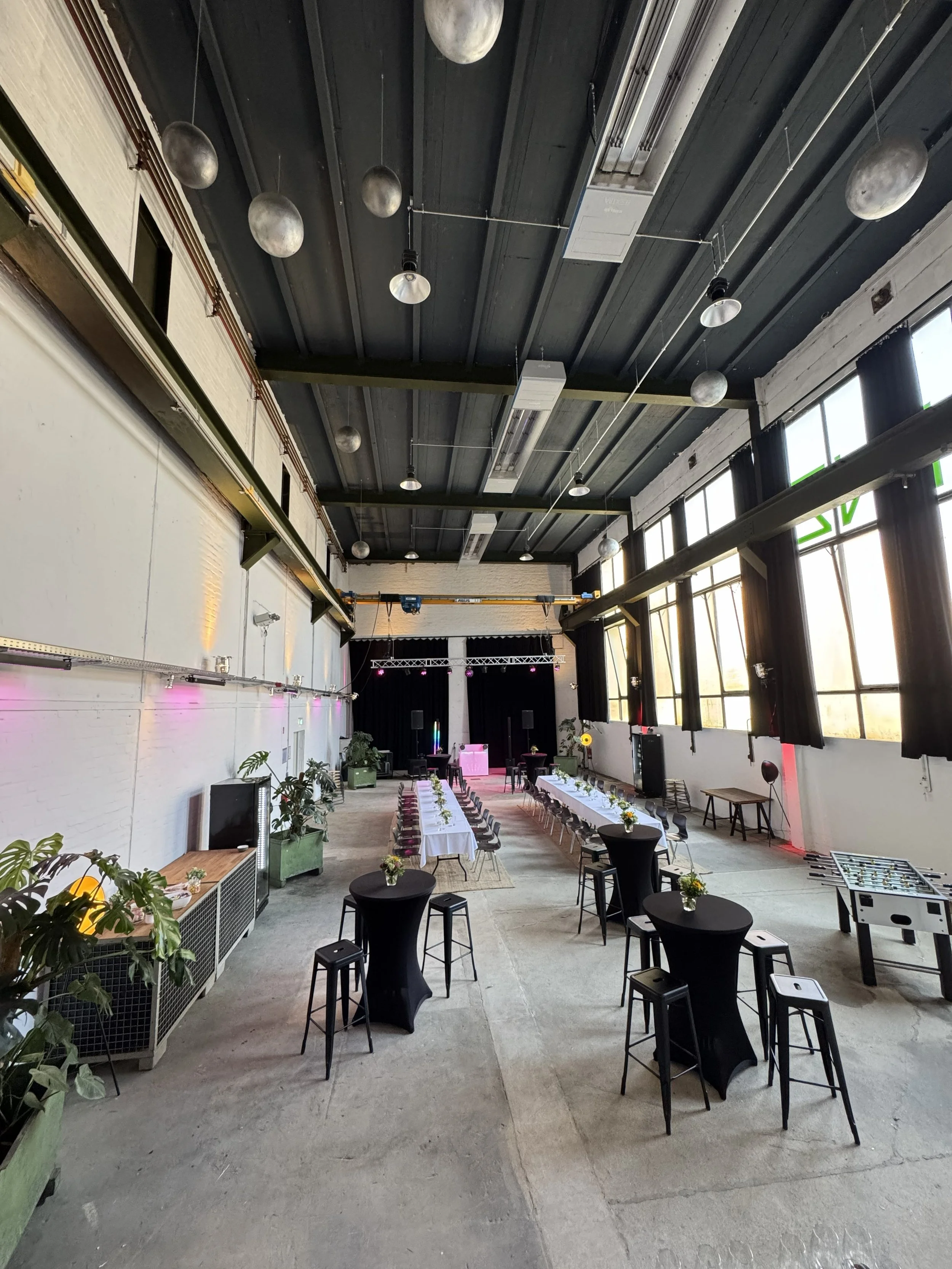 Großer Veranstaltungsraum mit hohen Decken, schwarzen Vorhängen, mehreren Tischen, die mit weißen Tüchern und kleinen Blumenvasen dekoriert sind, einigen Stehtischen, Pflanzen, und einem Showbühnenbereich mit Beleuchtung.