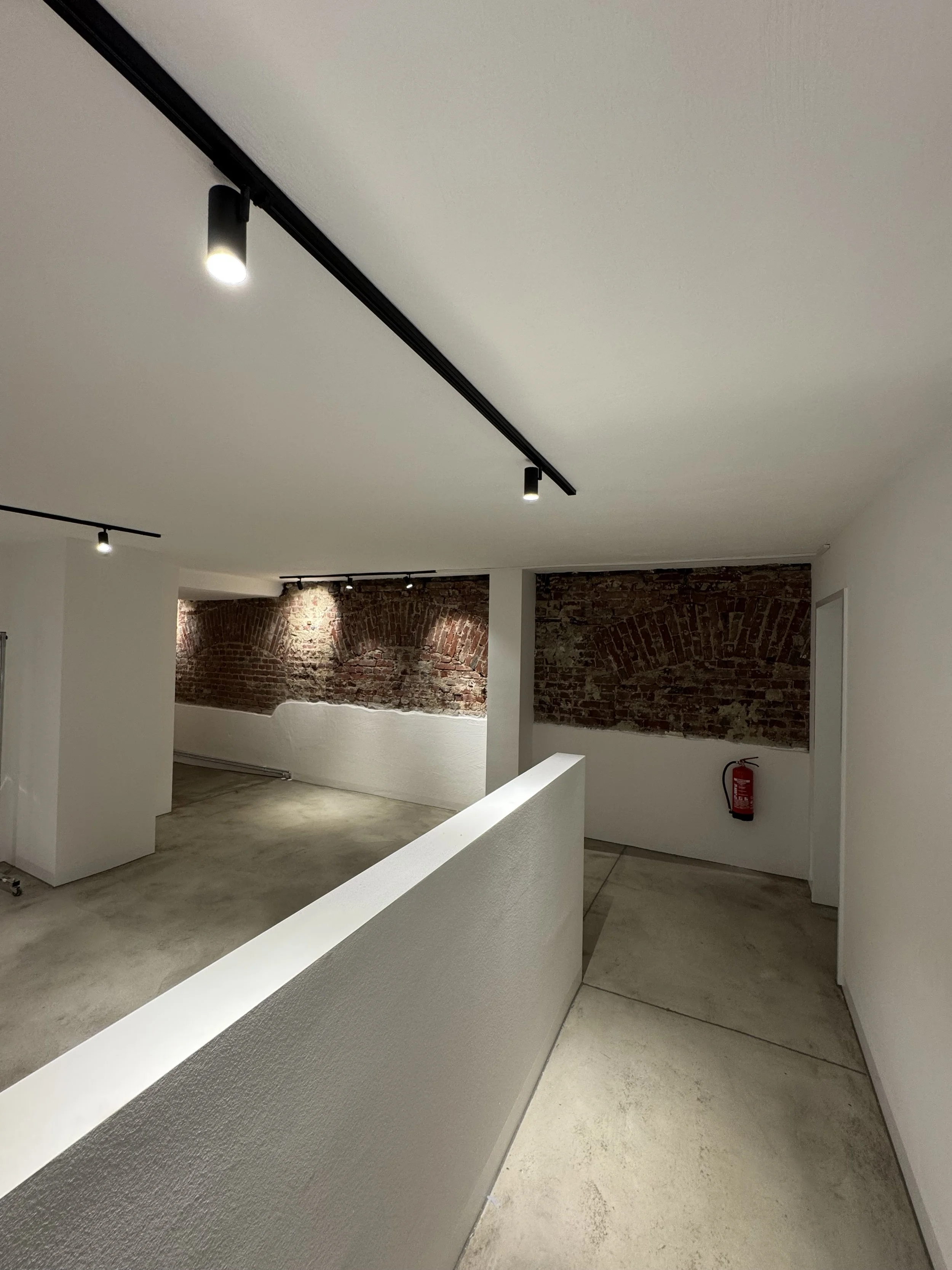 Innenraum einer modernen, offenen Raumgestaltung mit weißen Wänden, einer Trennwand und einer Wand mit Ziegelmauer im Hintergrund. Es gibt Deckenstrahler, eine Feuerlöscher box, einen weißen Handlauf und eine Betonbodenoberfläche.