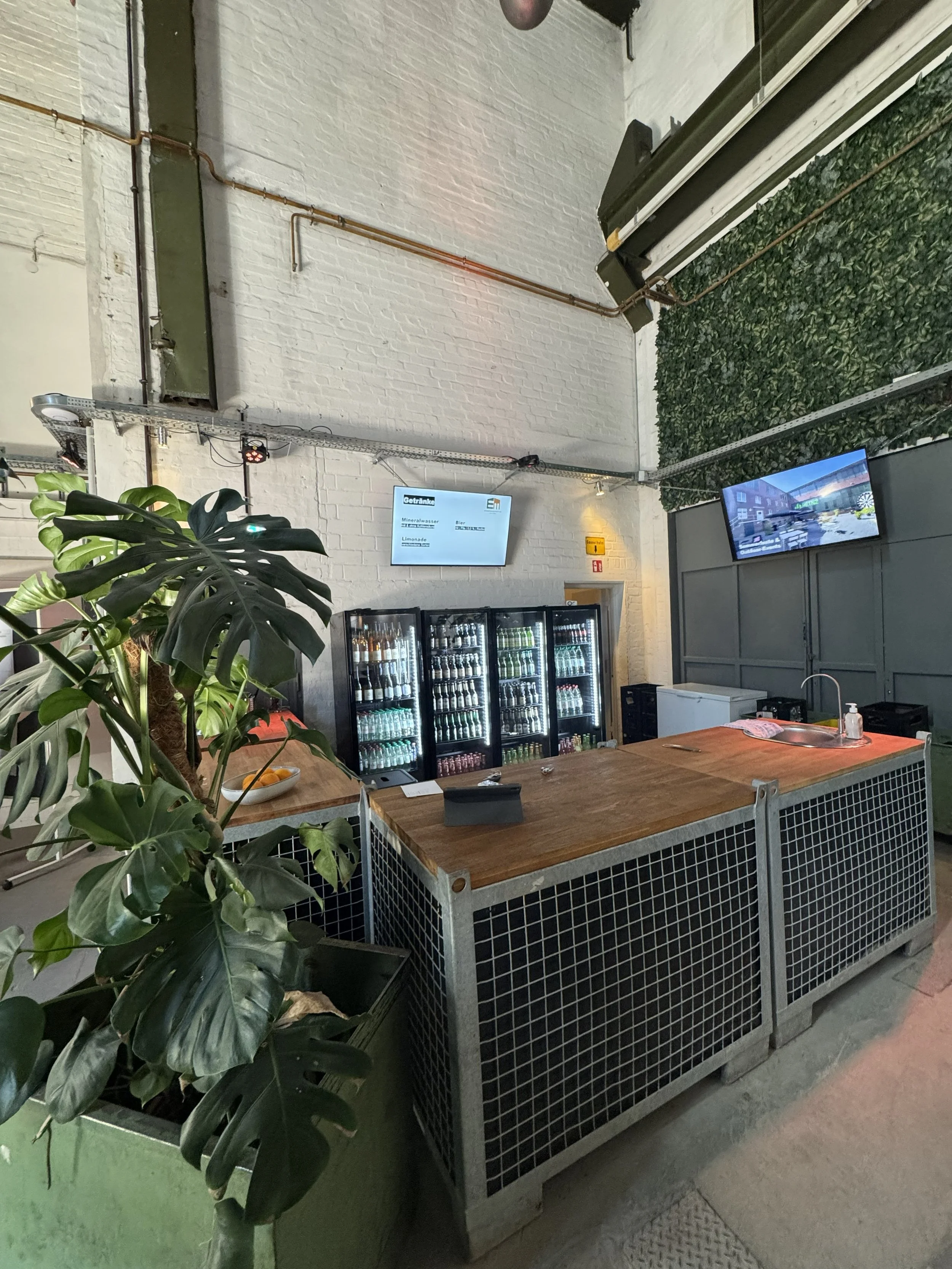 Ein Getränkeautomat mit verschiedenen Flaschen und Gläsern davor. In der Ecke eine große Zimmerpflanze, im Hintergrund ein weiß verputzter Ziegelsteinwand mit zwei Bildschirmen, einem größeren und einem kleineren, auf denen eine Innenaufnahme des Raums zu sehen ist.