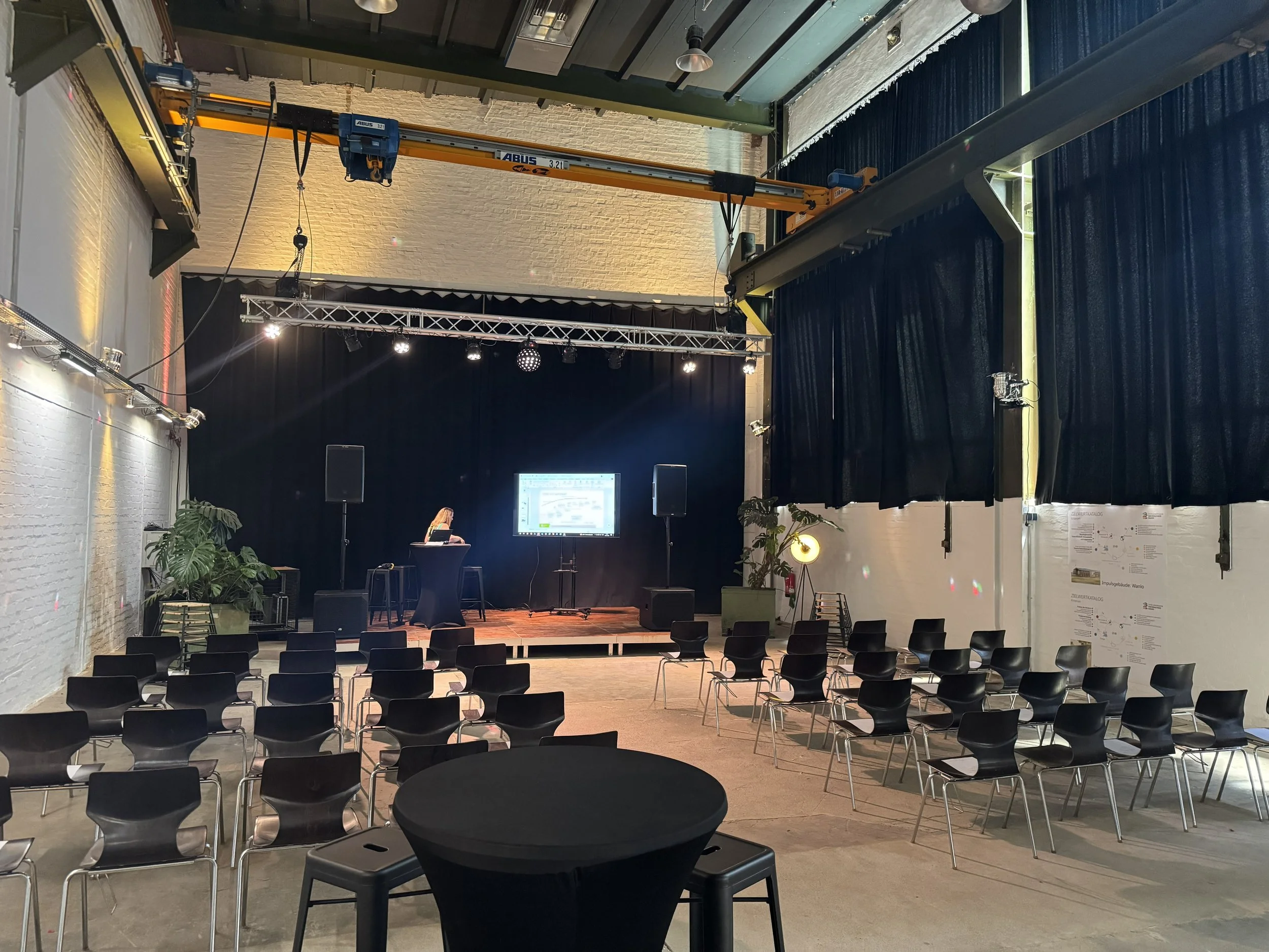 Ein Veranstaltungsraum mit einer Bühne, Stühlen im Saal, einer großen Leinwand, einem Podium mit Person, die einen Vortrag hält, und technischen Ausrüstungen wie Lautsprechern und Beleuchtung.
